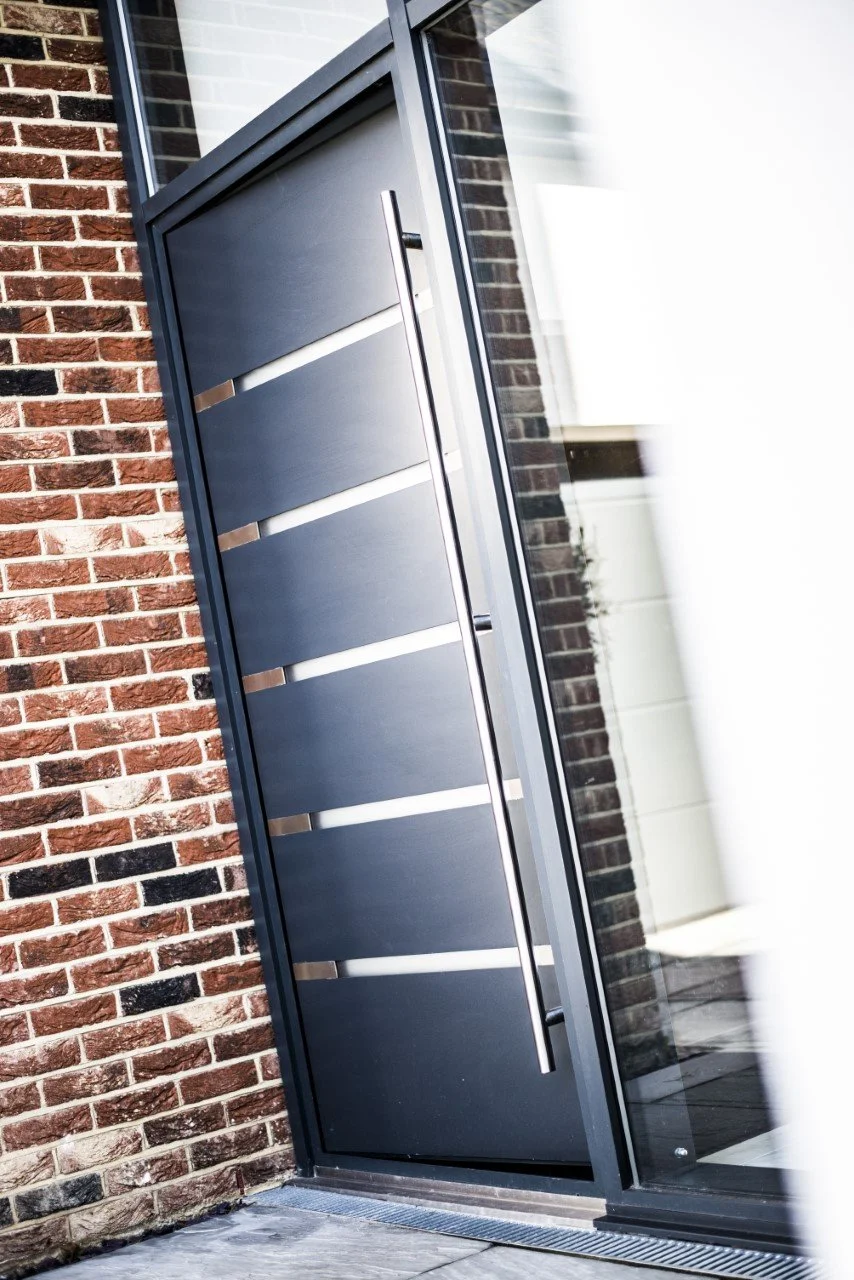 Modern black metal mailbox installed beside a brick wall and glass door at an entrance.