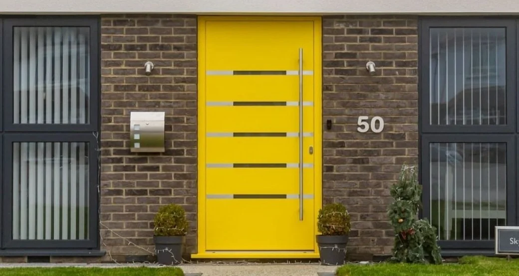 Modern house with yellow front door, brick exterior, two windows with vertical blinds, potted plants, and house number 50.