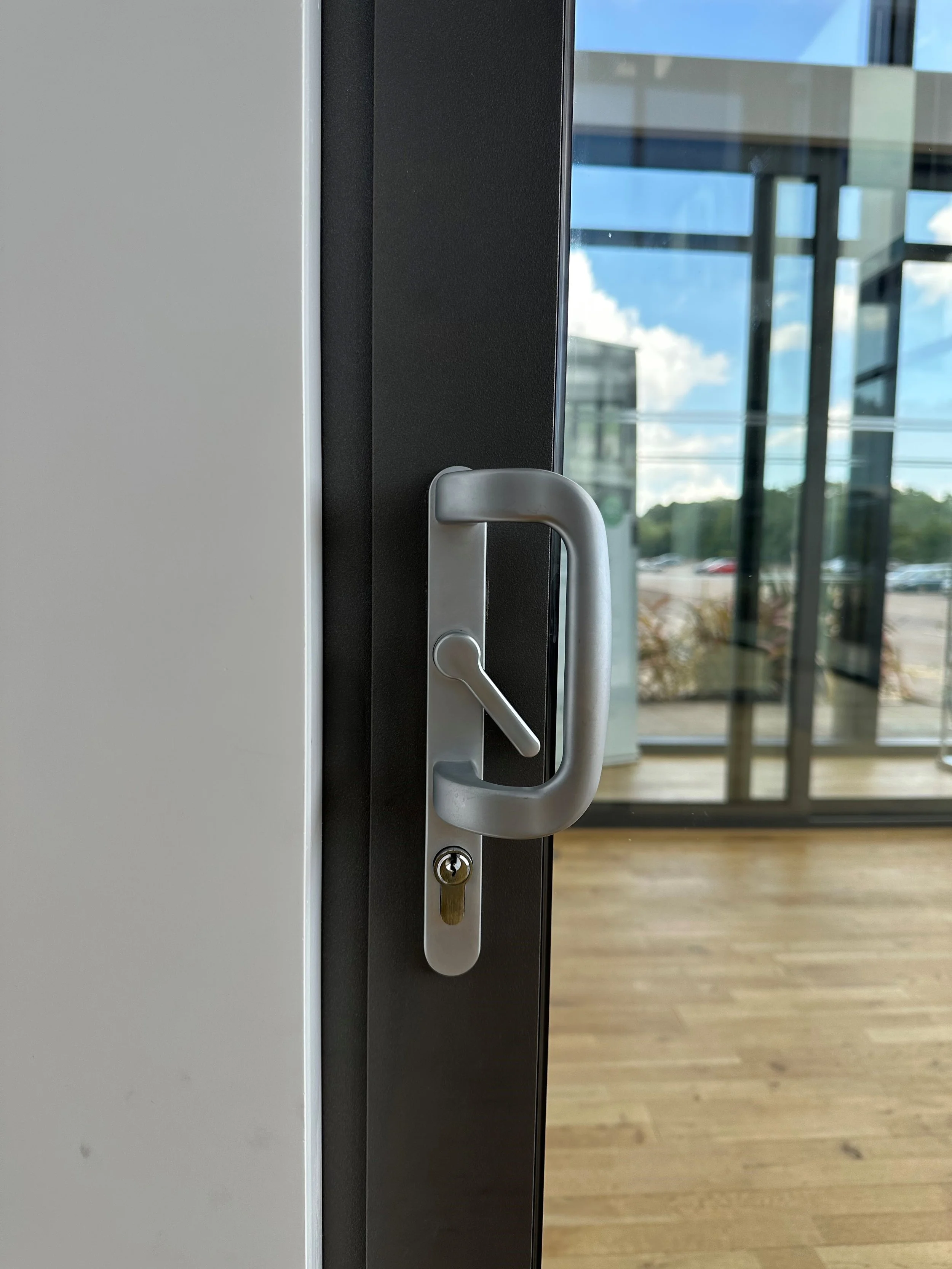 Close-up of a silver door handle with a keyhole, on a glass door, revealing an interior space with wooden floors and large glass windows showing a cloudy sky outside.
