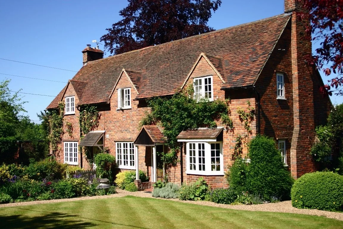A two-story brick house with multiple white-framed windows, surrounded by a well-maintained garden with green grass, shrubs, and trees, under a clear blue sky.