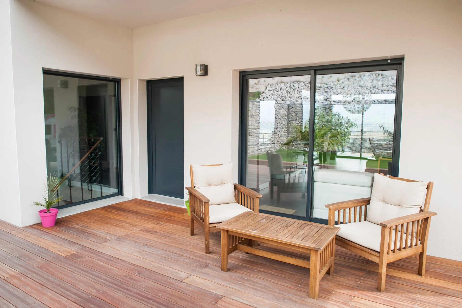 A balcony with wooden flooring, two wooden chairs with white cushions, a matching wooden table, a potted plant in a pink pot, and glass sliding doors leading to an indoor space.