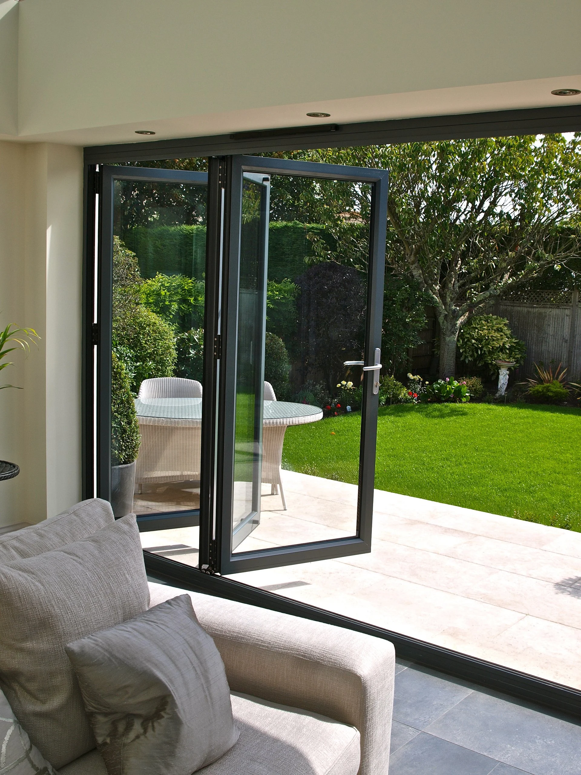Open glass sliding door leading to a backyard with green grass, plants, and trees, seen from inside a living room.