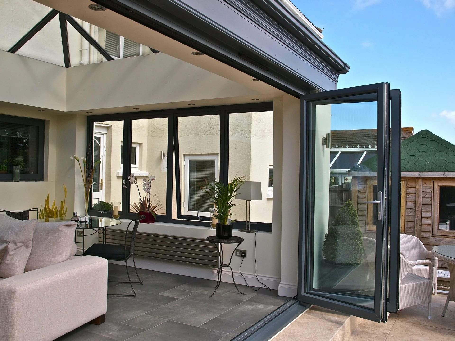 An indoor patio with a sofa, chairs, and plants near large windows and an open glass door leading outside to a patio with a wooden shed and garden.