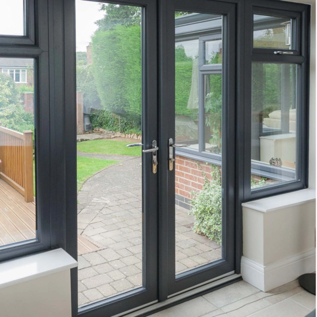 View through a black-framed glass door leading to a backyard with paving stones, green grass, and trimmed hedge.