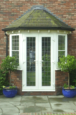 Double glass doors with white frames and grid panes, set in a brick wall, with two potted green plants in blue pots on either side and a paved patio in front.