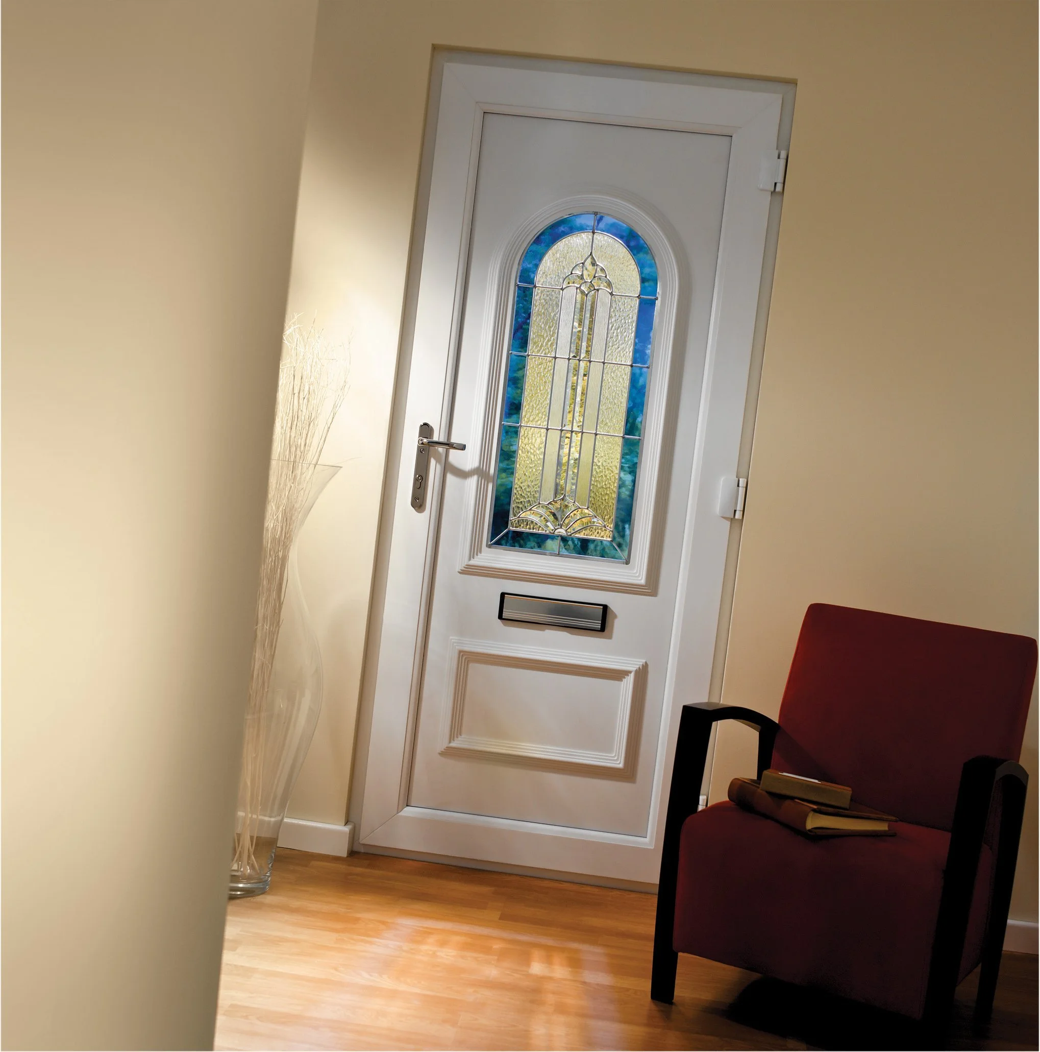 An interior view of a front door with a decorative stained glass window, a red armchair with books on it, and a tall vase with dried decorative grass in the corner.