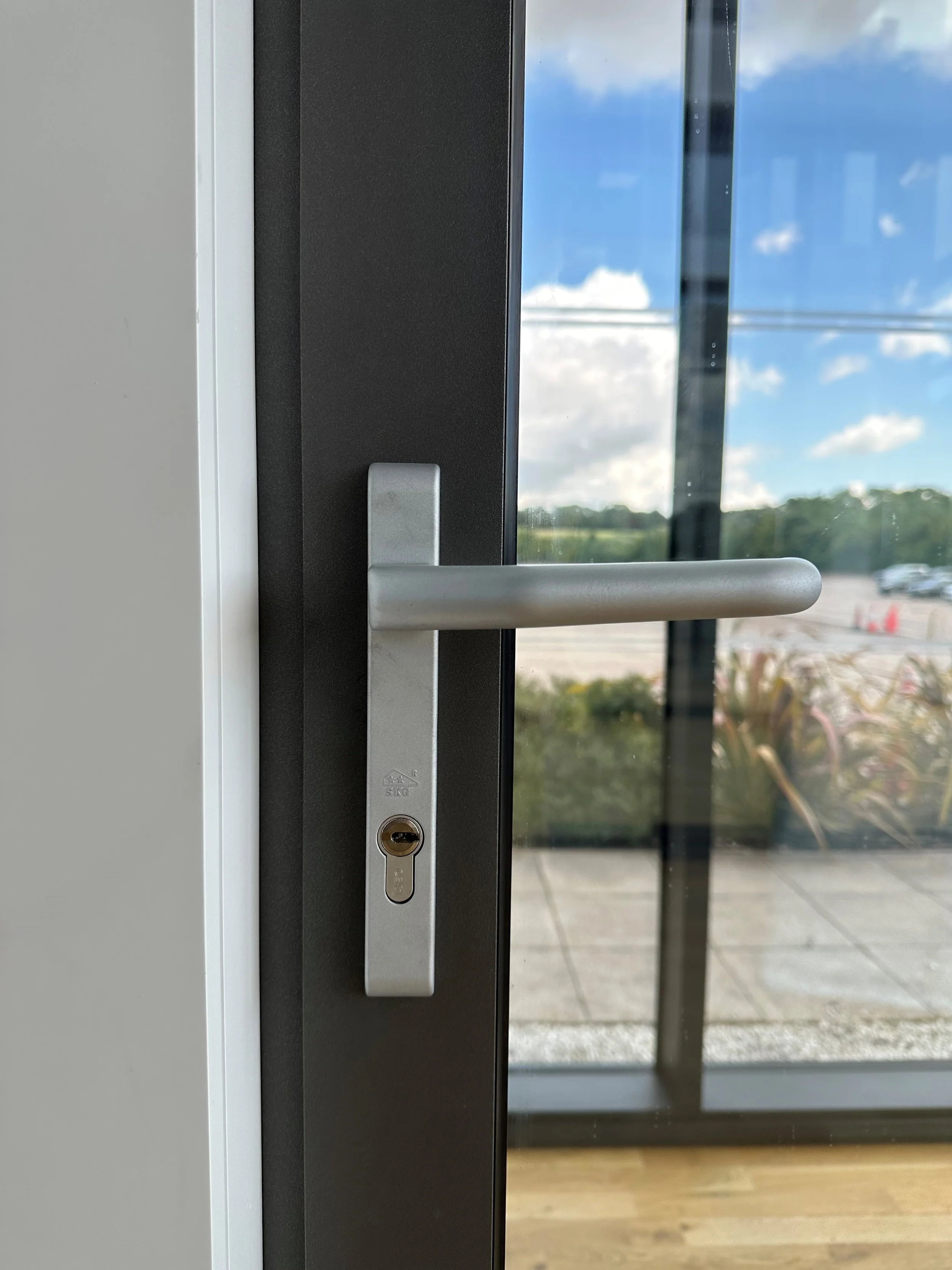 Close-up of a silver door handle with a keyhole on a glass door, showing an outdoor parking lot and blue sky with white clouds through the glass.