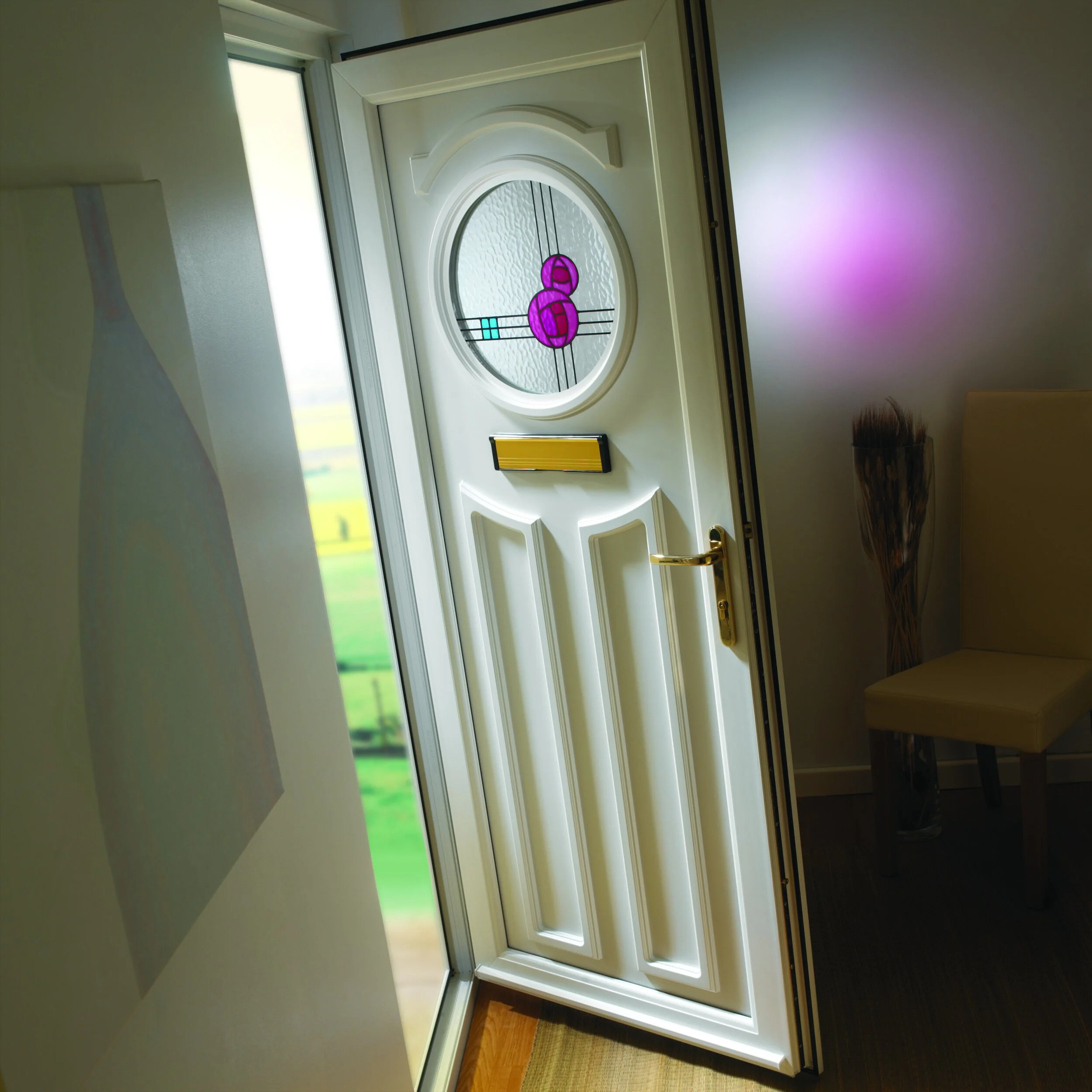 Close-up of a white front door with decorative stained glass window featuring pink and blue patterns, gold handle, and mail slot, open to reveal a view of green fields outside.