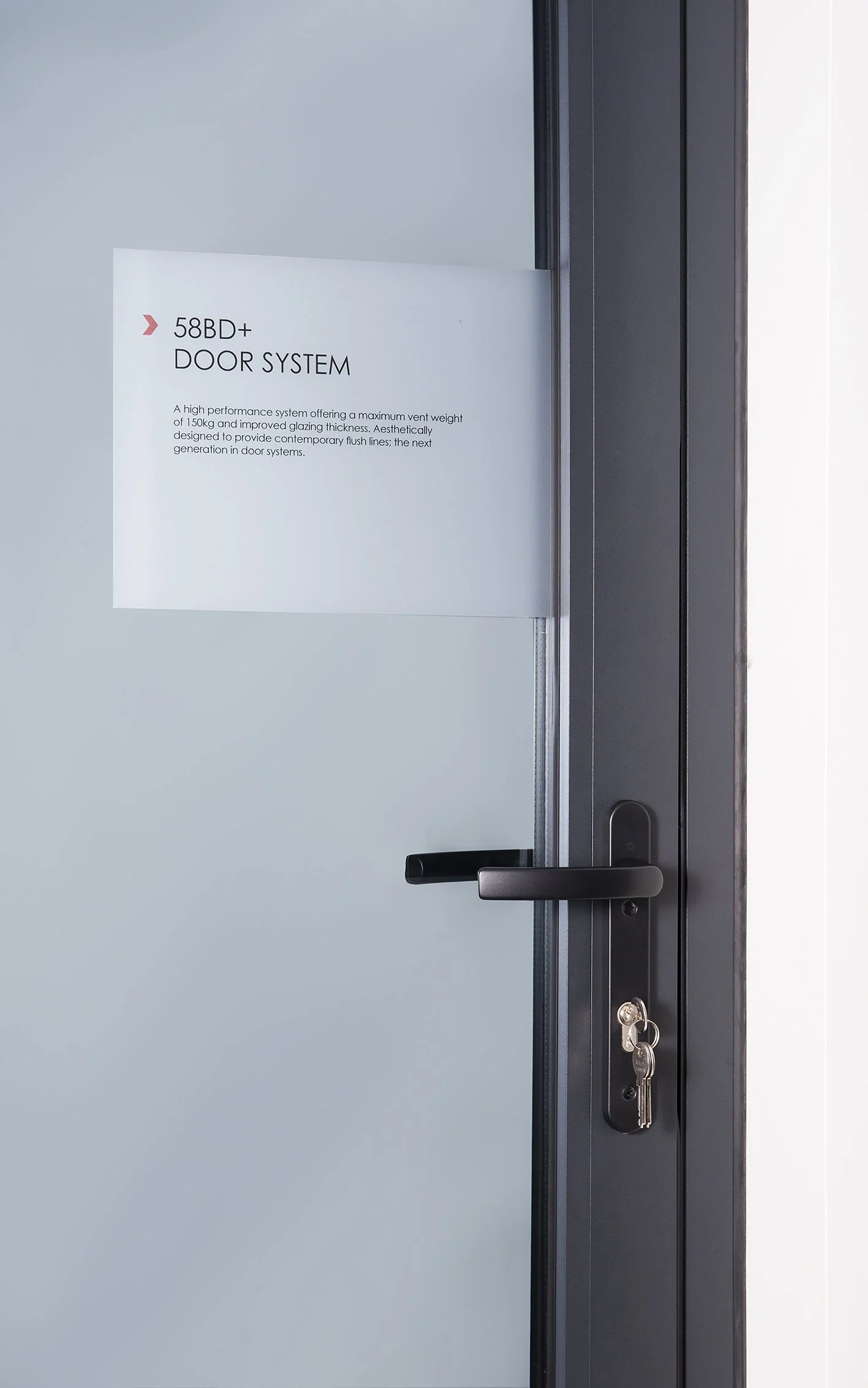 Glass door with a black handle and a key in the lock, with a white sign on it that says '58BD+ DOOR SYSTEM' and descriptive text about a high-performance door system.