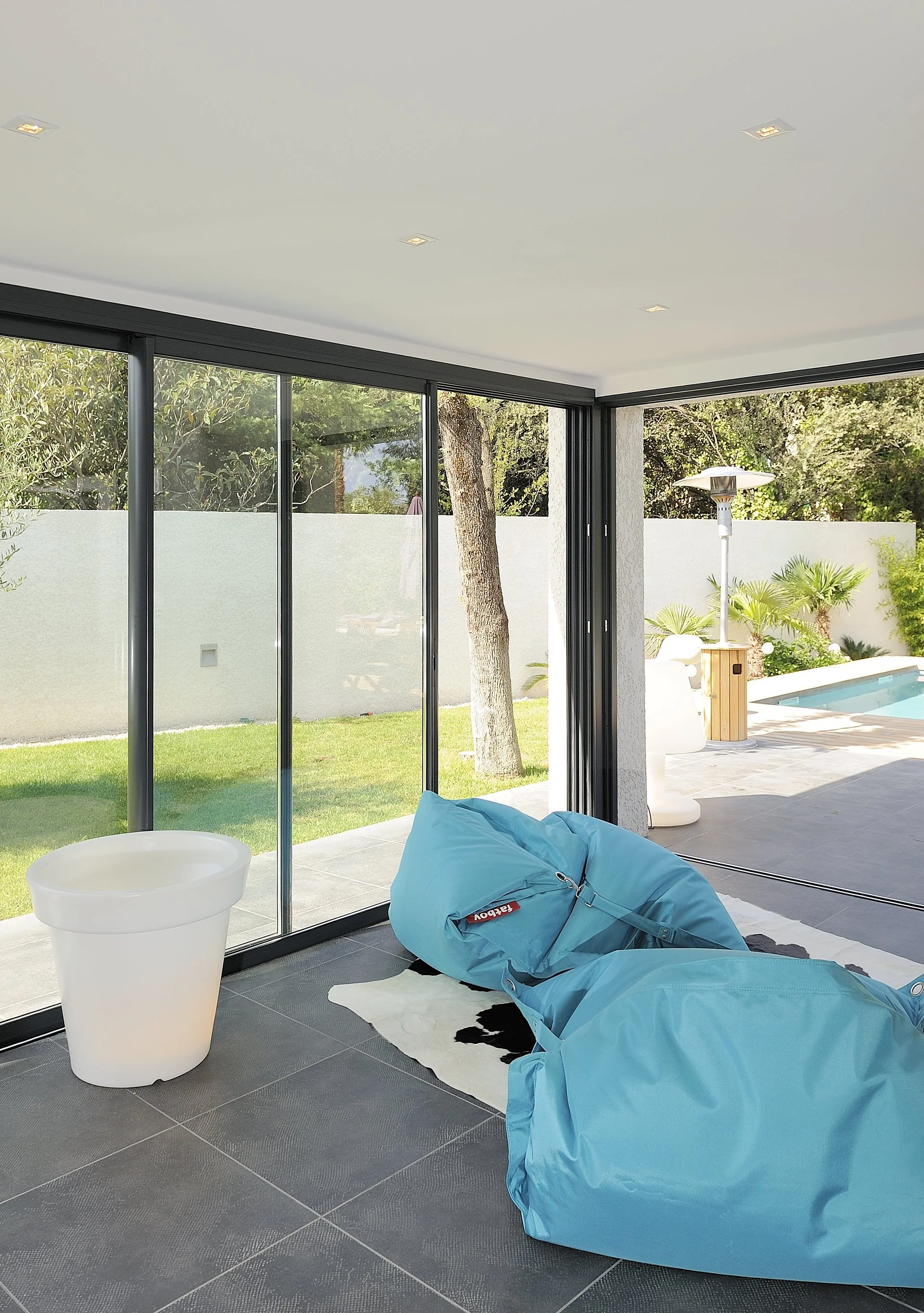 Modern living room with large glass sliding doors opening to a backyard with a tree, grass, and a pool; includes a blue bean bag chair, a white planter, and a cowhide rug.