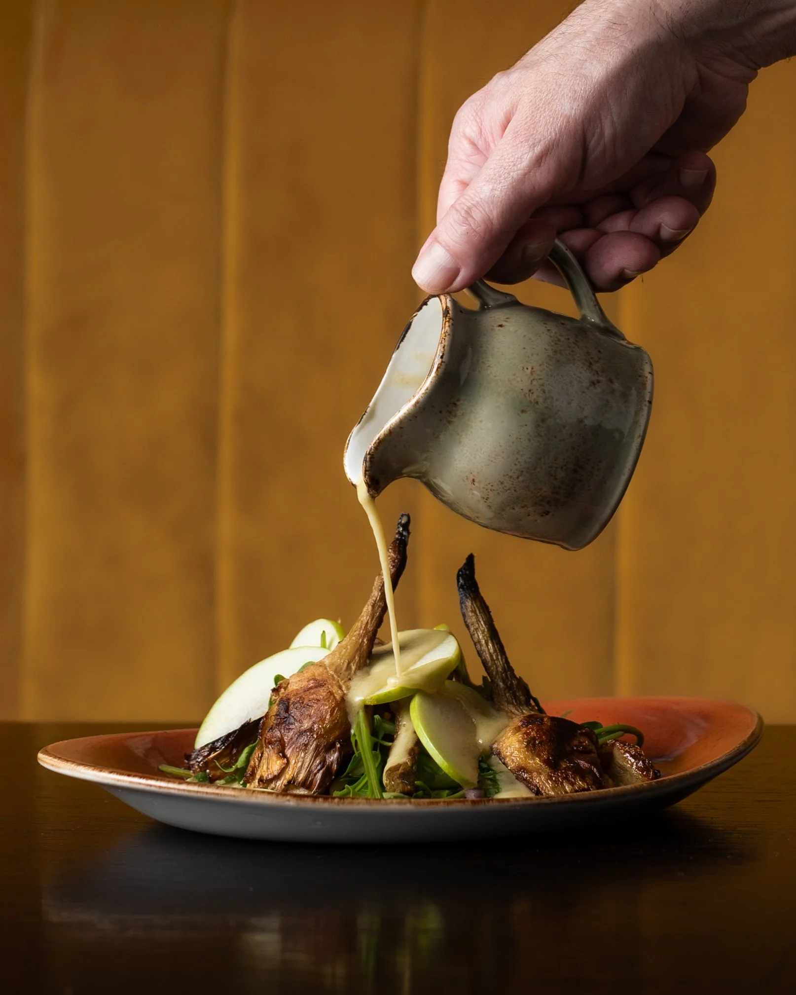 Hand pouring sauce over roasted quail served with sliced apples and greens on a plate.