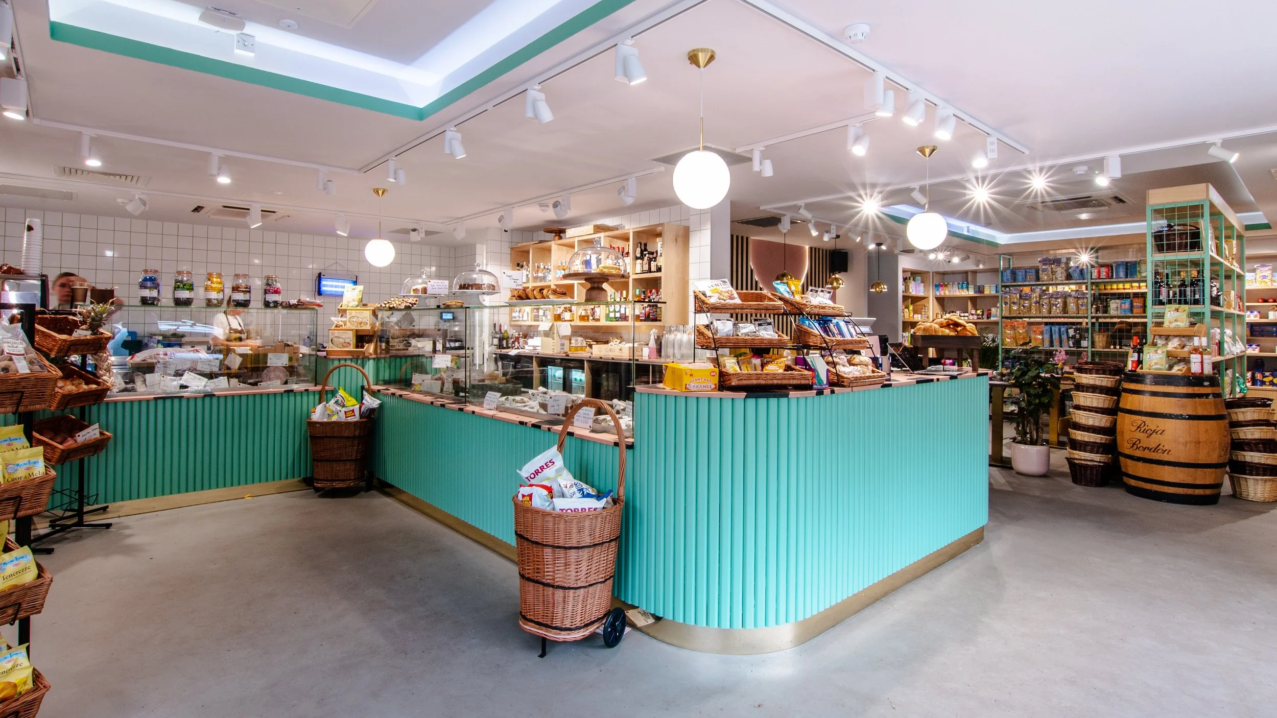 Interior of a modern grocery store with teal-colored counter, various snacks and baked goods, shelves with products, and decorative barrels.