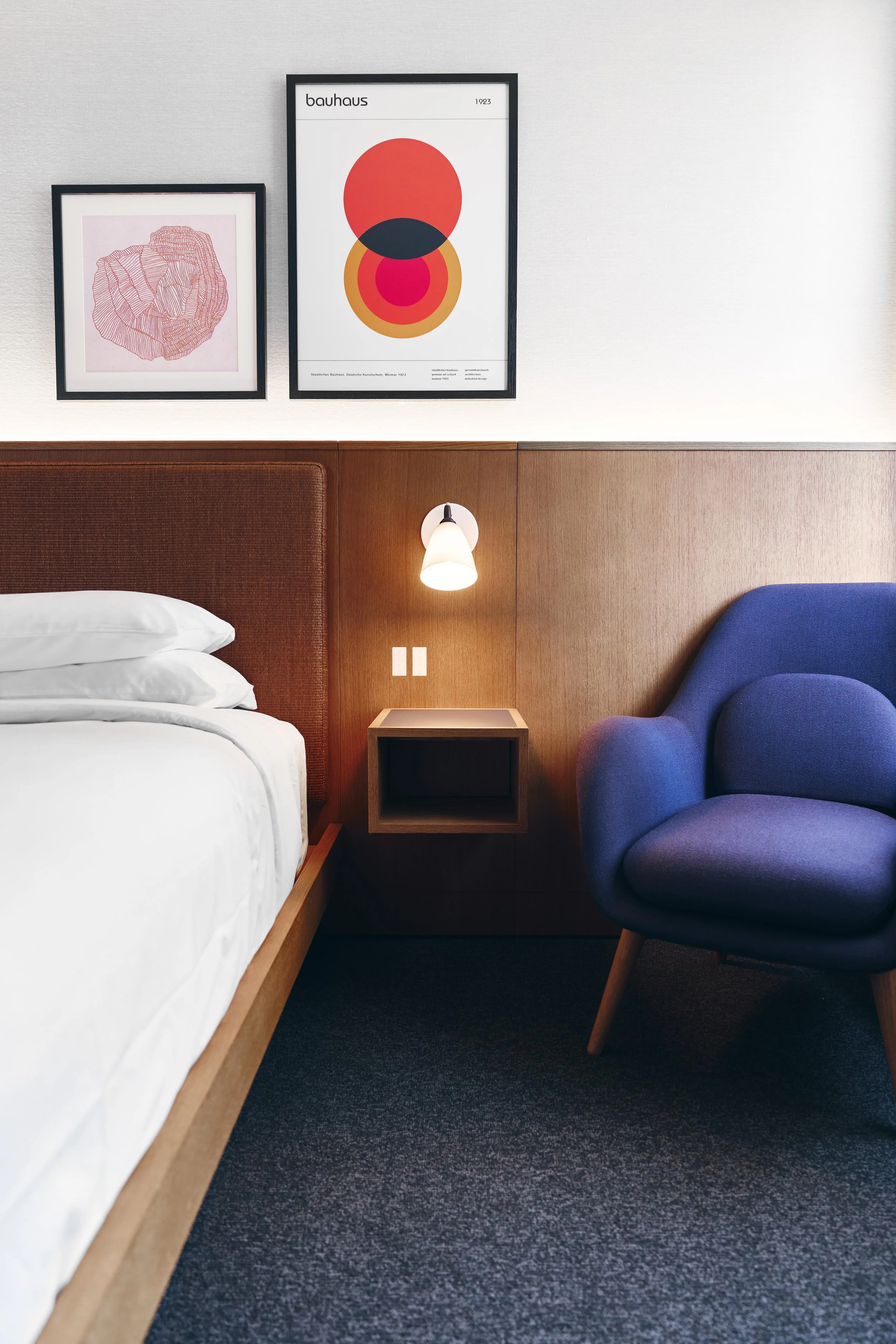 Hotel bedroom with a white bed, dark wood headboard, blue armchair, and wall-mounted light fixture. Artwork with abstract and geometric prints hang on the white wall.