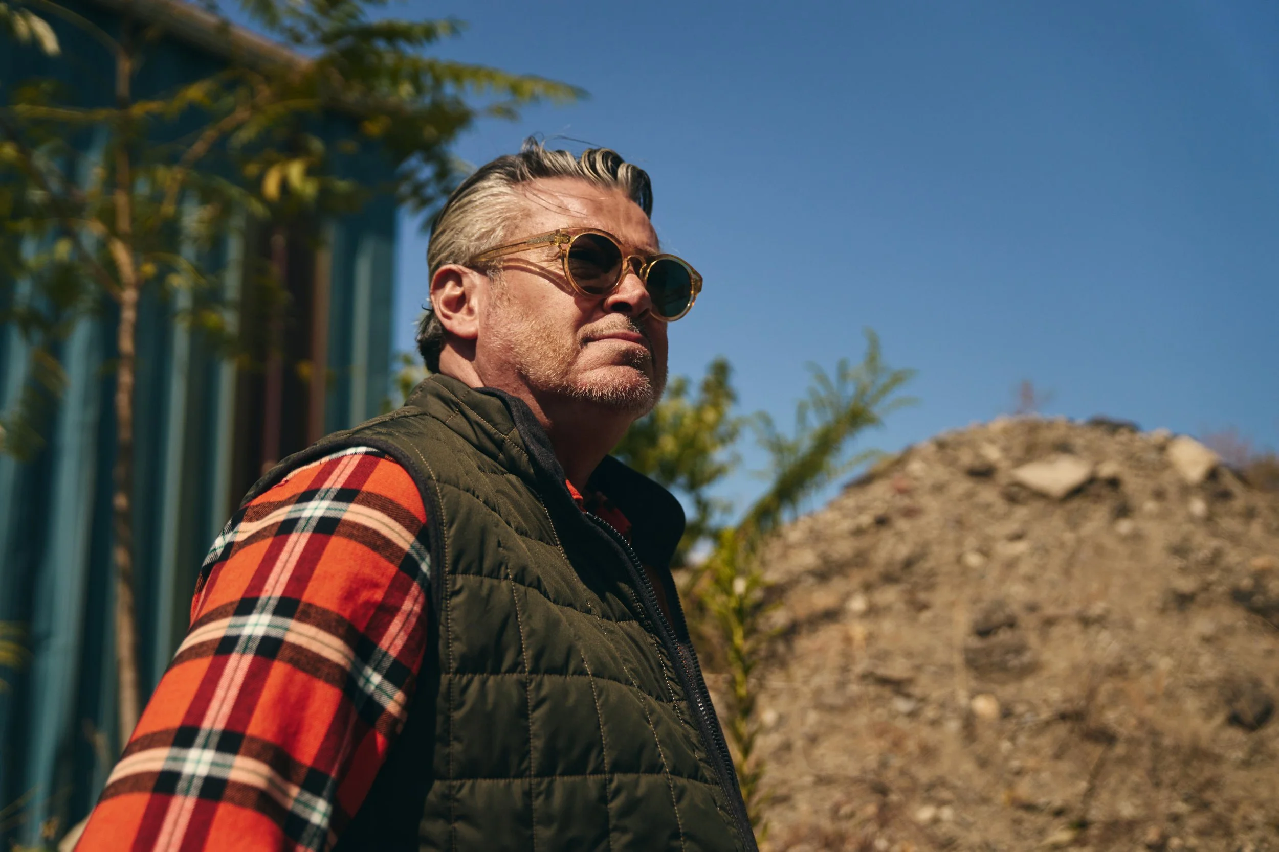 A man with gray hair, sunglasses, and a beard stands outdoors against a clear blue sky, wearing a plaid shirt and a quilted green vest, with a mound of dirt or rocks behind him.