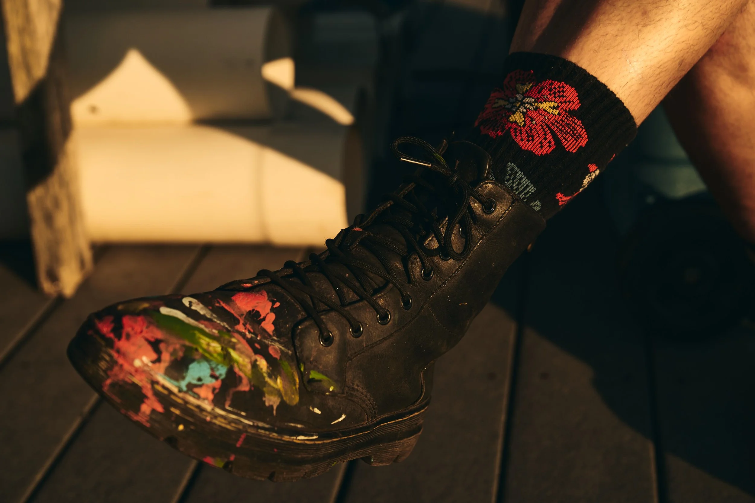 A close-up of a black leather boot with multicolored paint splatters, worn with a black sock featuring a red hibiscus flower design. The background shows a dimly lit outdoor setting with wooden flooring and a box with a partial shadow.