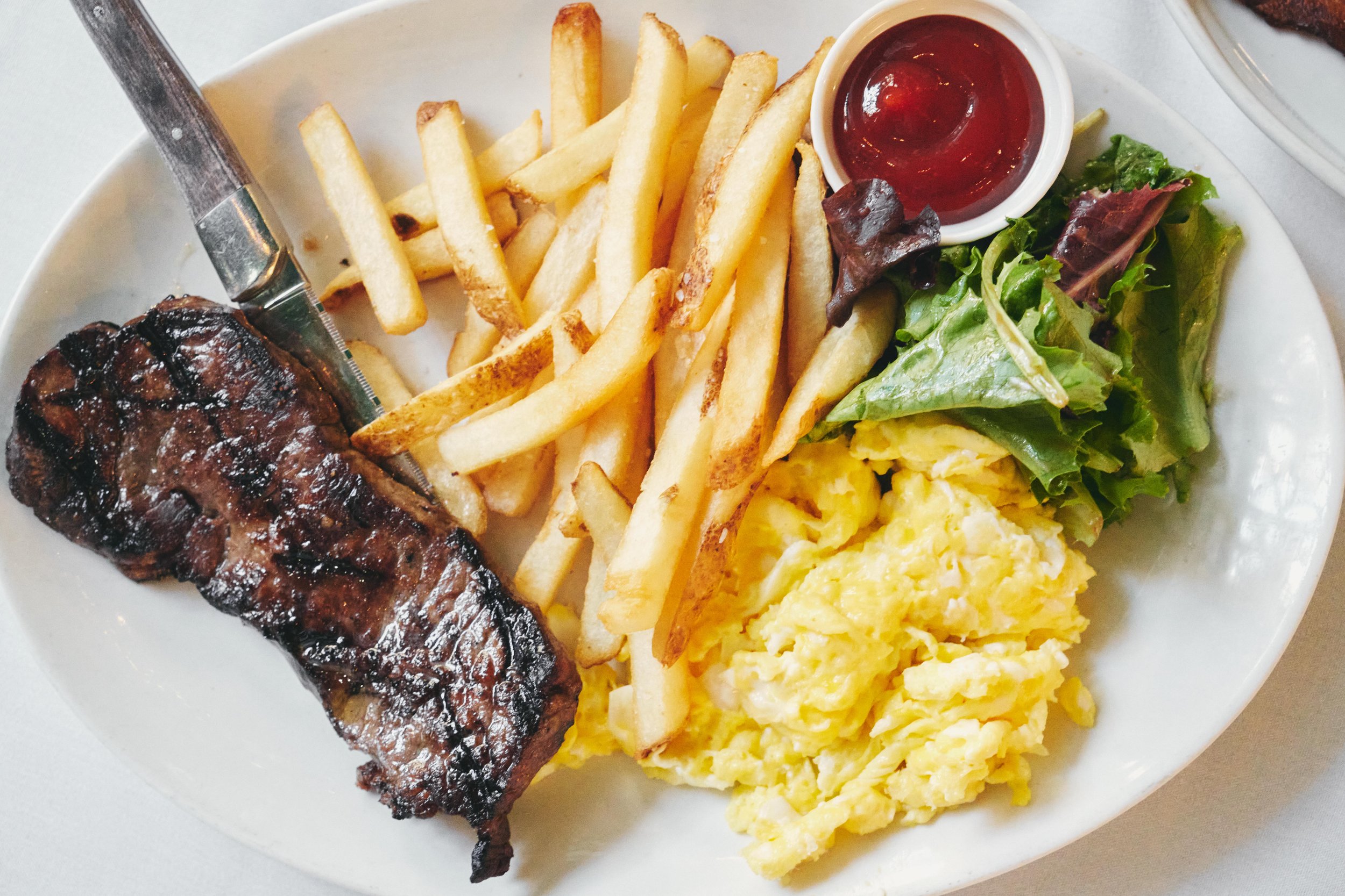 Plate of grilled steak, French fries, scrambled eggs, mixed greens with lettuce, and a small cup of ketchup.