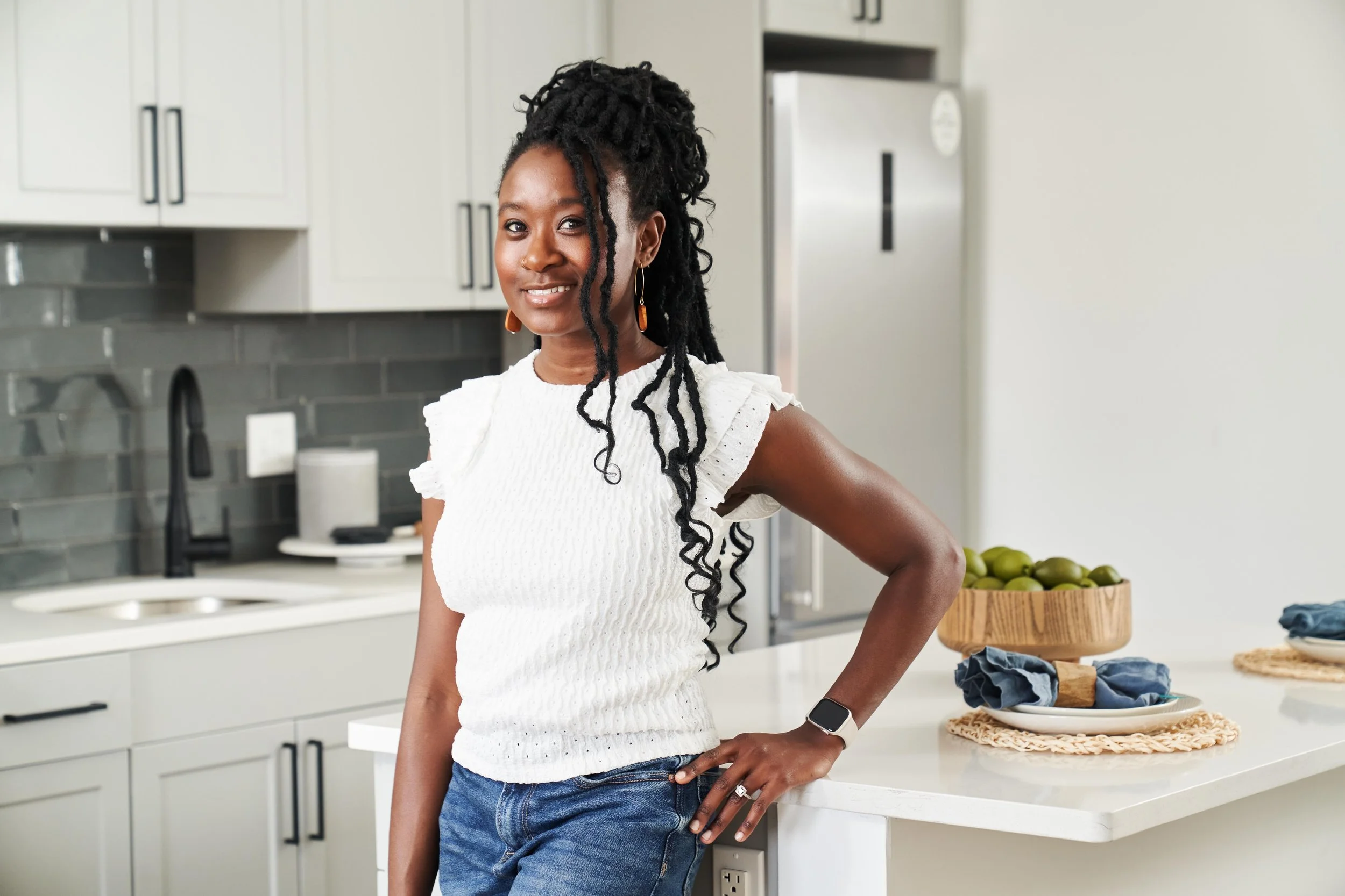 A woman with dark, curly hair styled in dreadlocks is standing in a modern kitchen, smiling at the camera. She is wearing a white sleeveless top with ruffled shoulders, blue jeans, and a smartwatch on her left wrist.