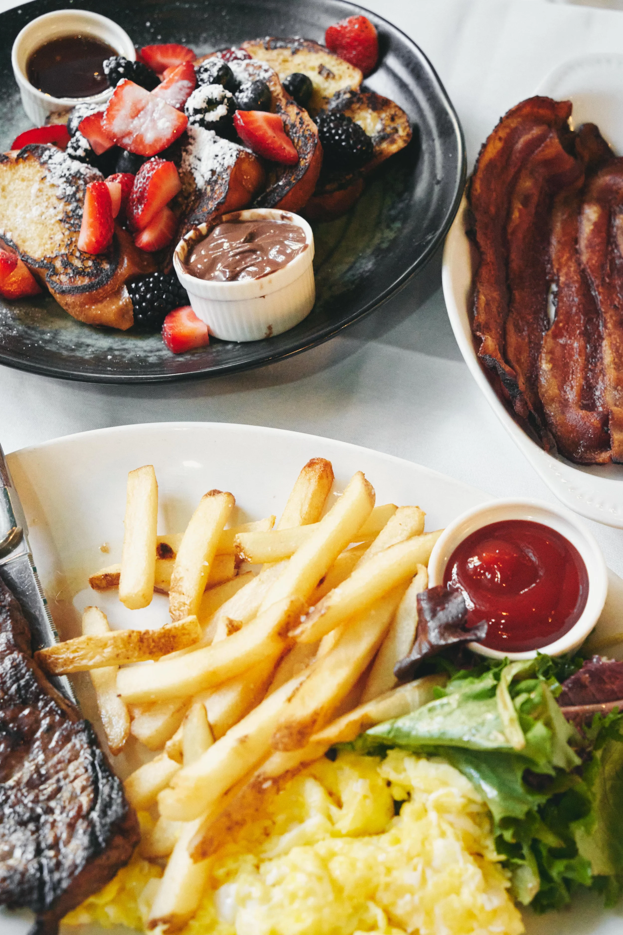 Breakfast with French toast topped with strawberries, blackberries, and powdered sugar, served with chocolate and syrup sauces; side of bacon; plate of French fries; scrambled eggs; salad with lettuce, and a small cup of ketchup.