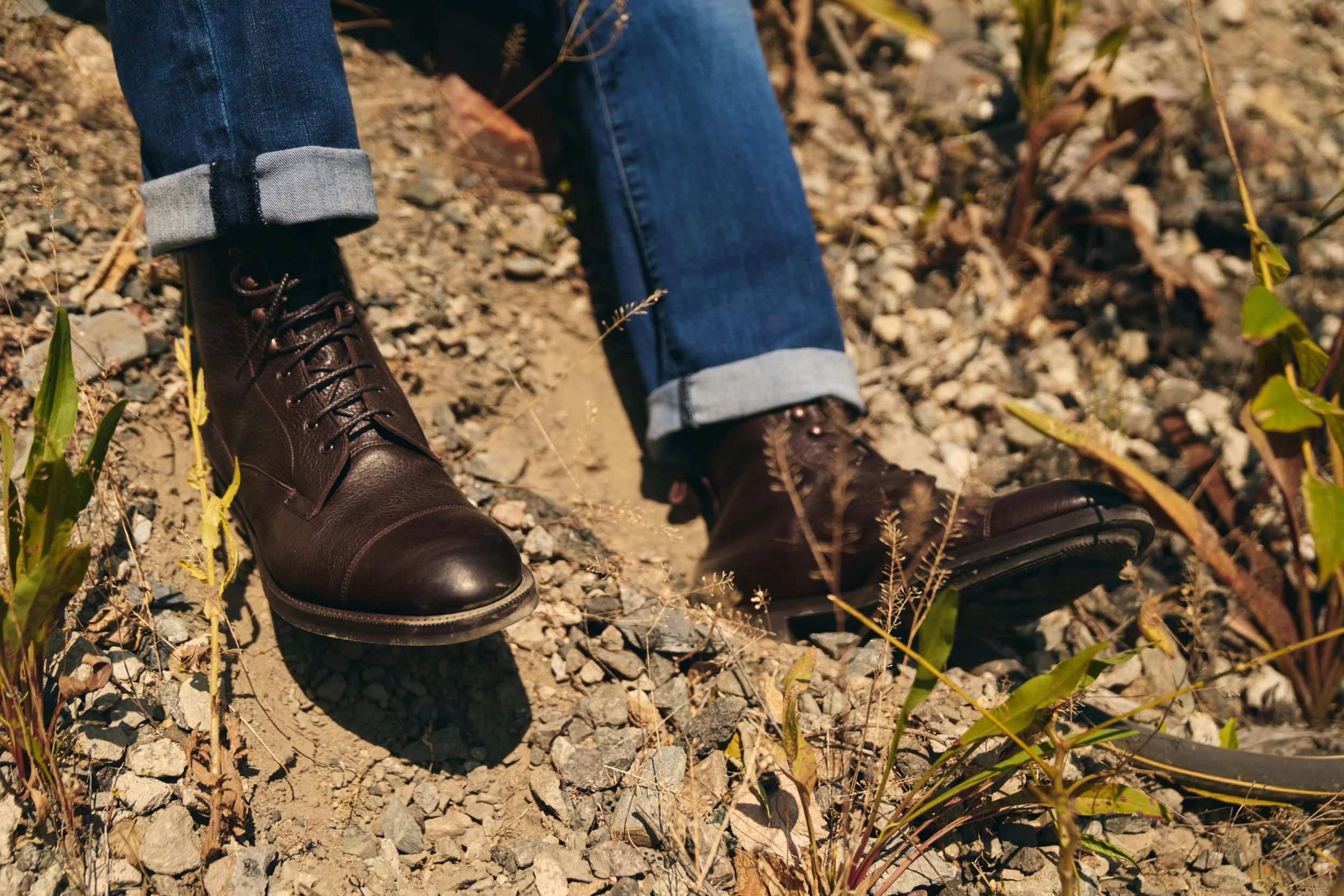 Person wearing dark brown leather boots and rolled-up blue jeans walking on rocky dirt ground with sparse green plants.
