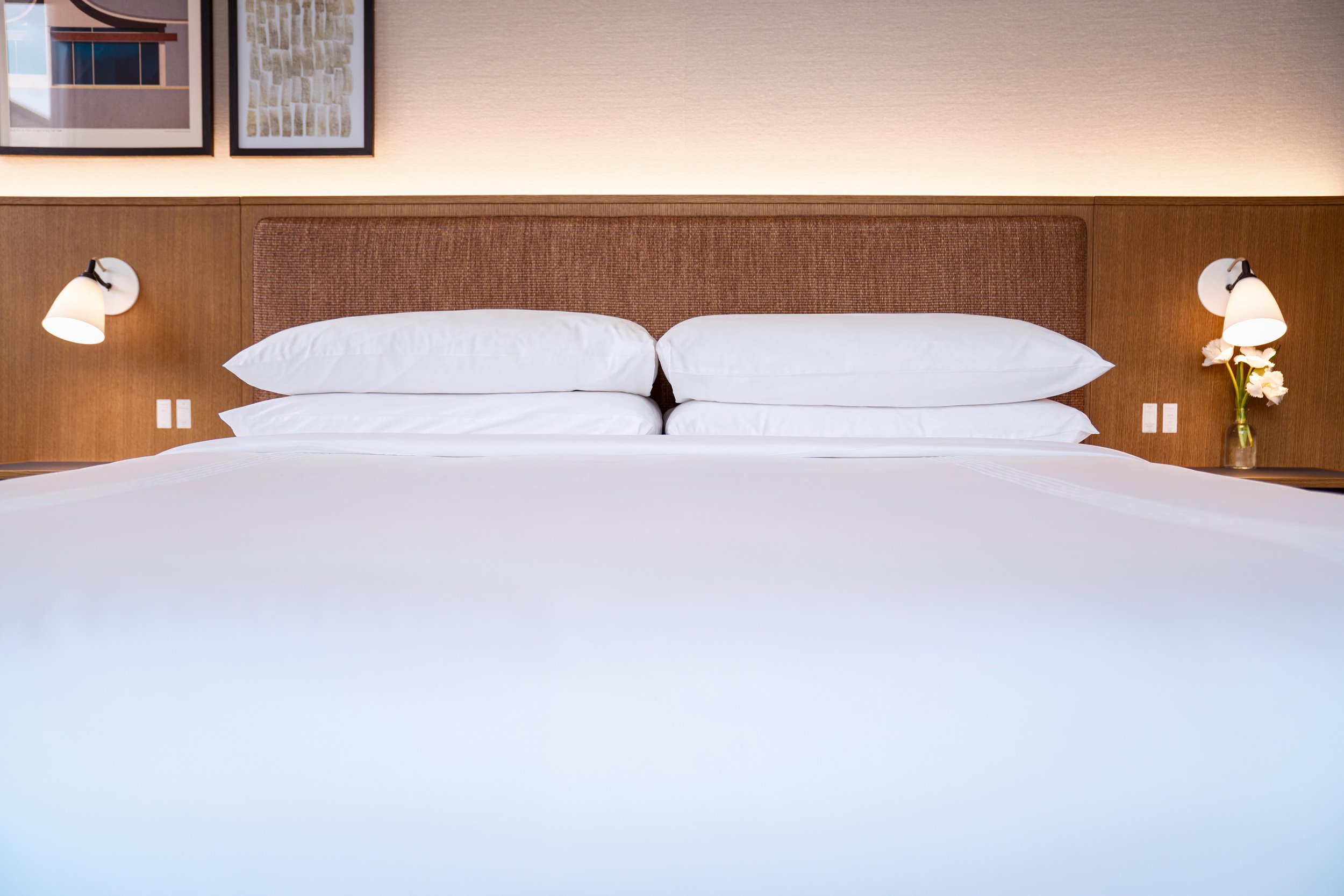 A neatly made bed with white pillows and bedding, a wooden headboard, two wall-mounted lamps, and framed wall art above the bed, with a soft light illuminating the room.