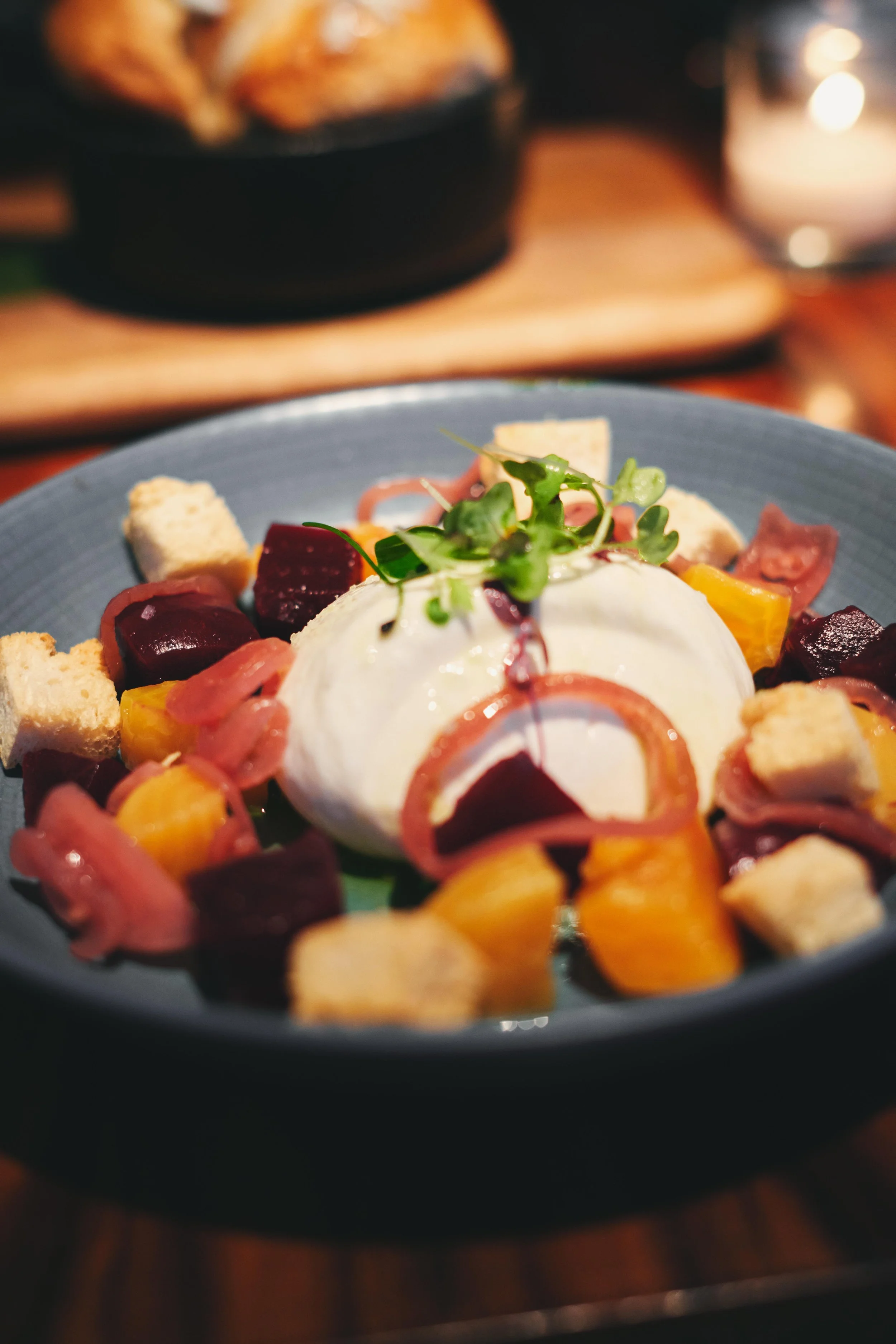 A black plate with a poached egg topped with microgreens, surrounded by diced yellow and beets, pickled onions, bread cubes, and garnished with herbs.