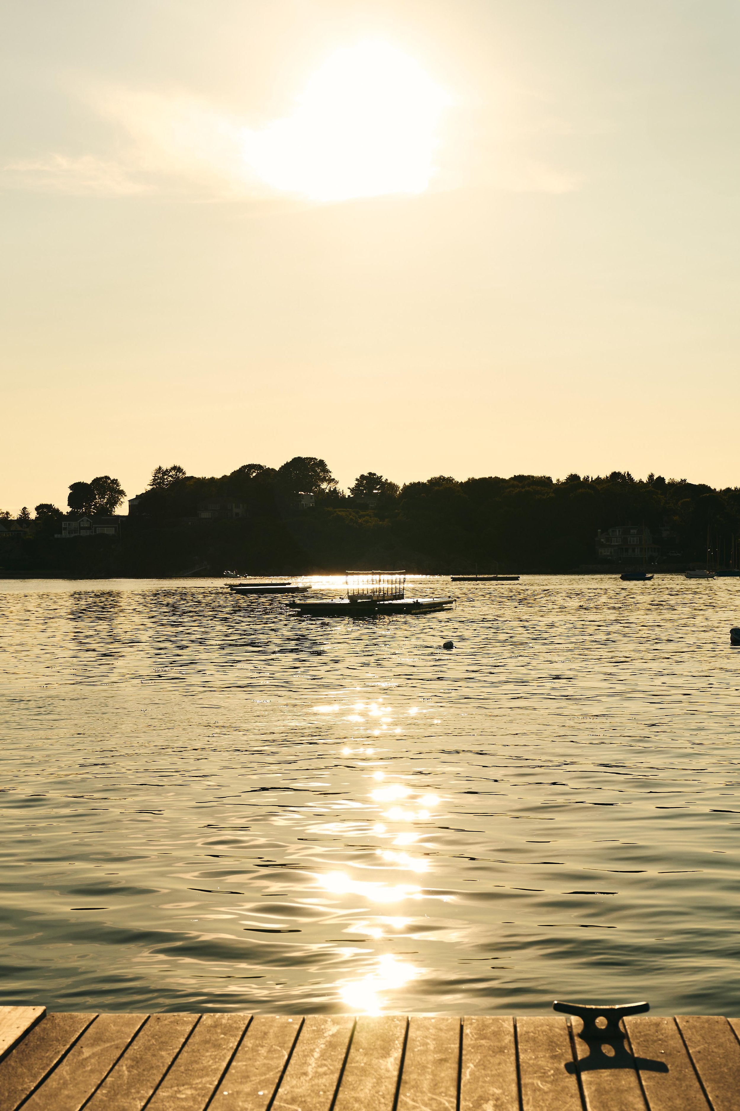 Sunset over a calm body of water with reflections on the surface, boats floating, and a wooden dock in the foreground.