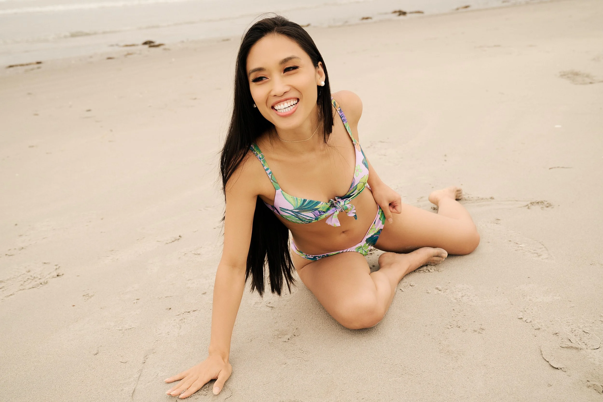 A smiling woman in a colorful bikini sitting on the sand at the beach.