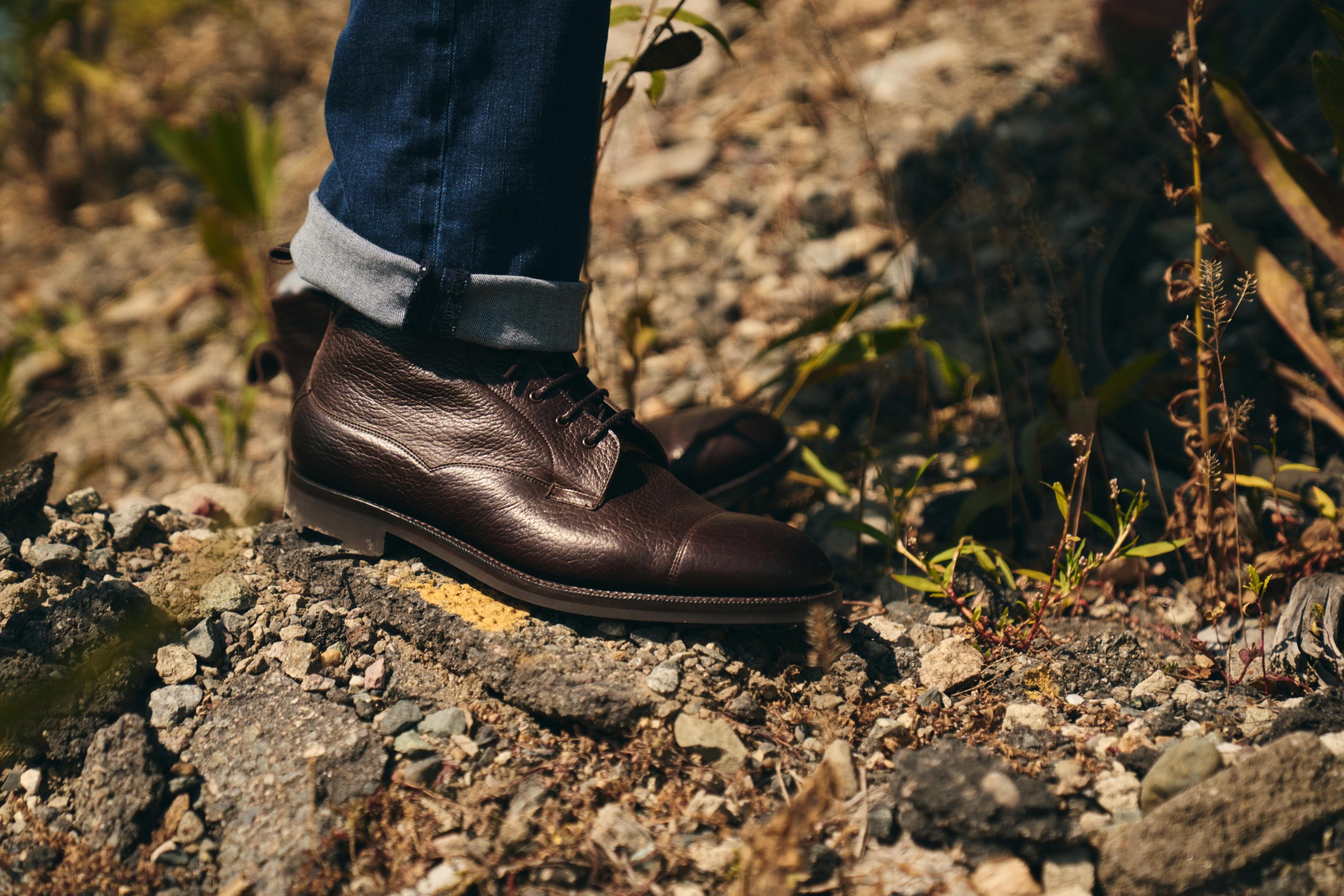 Close-up of a person's foot wearing a dark brown leather boot and blue jeans, standing on rocky, uneven ground surrounded by small plants and dry foliage.