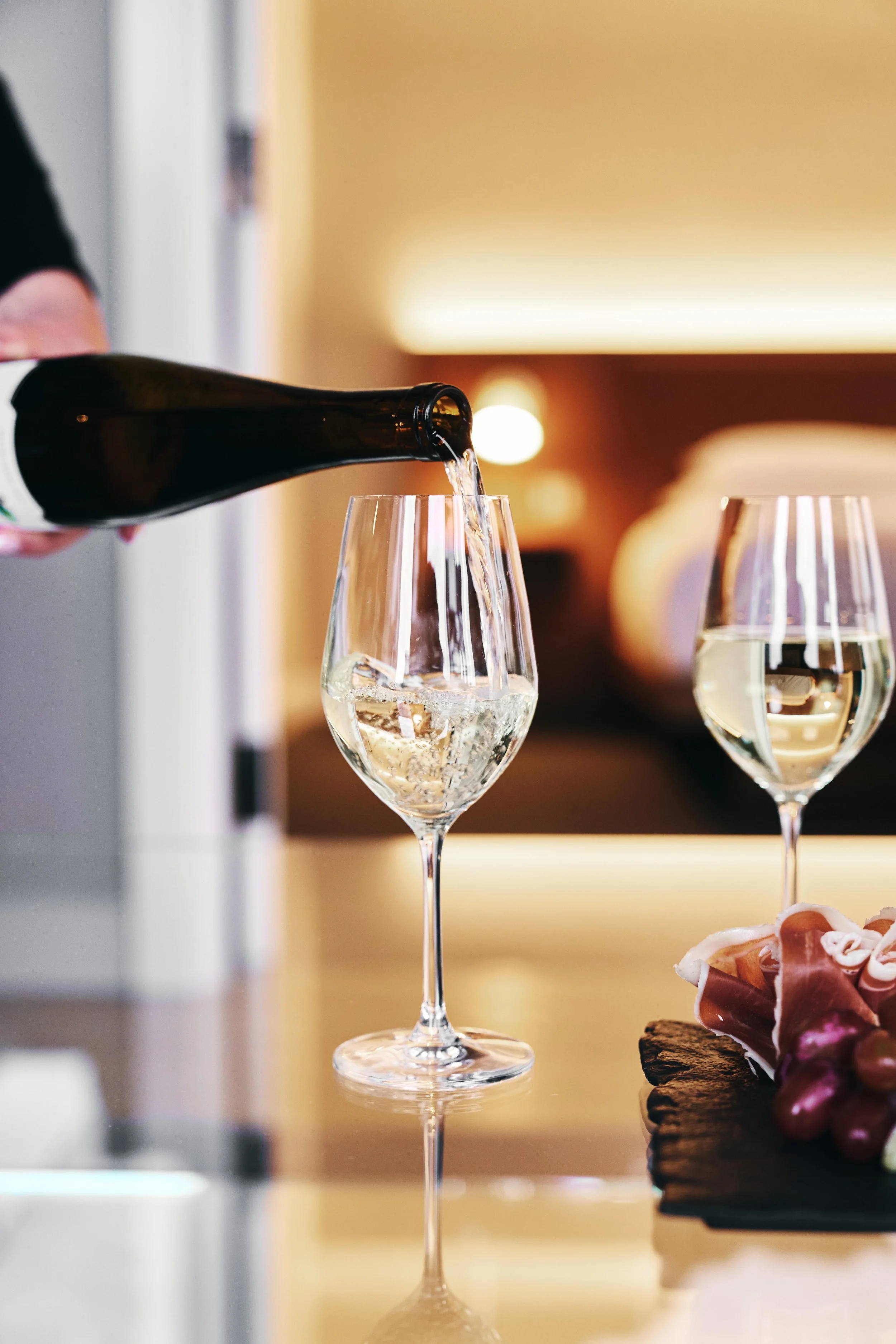 Person pouring champagne into a glass on a table, with another glass of champagne and a charcuterie board nearby.
