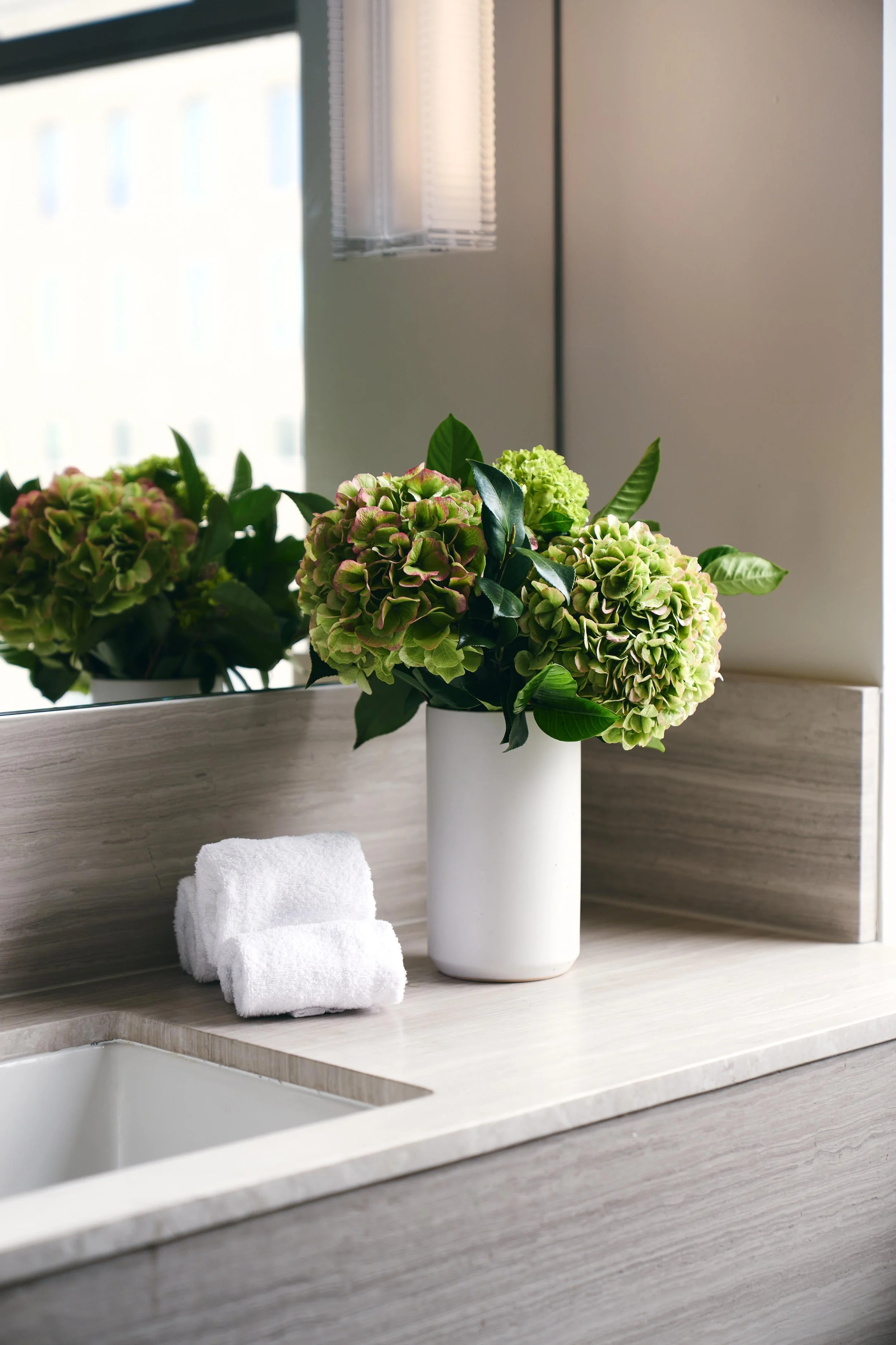 A white ceramic vase filled with green hydrangea flowers and green leaves on a light-colored countertop, with a mirror reflecting similar flowers and a window in the background.