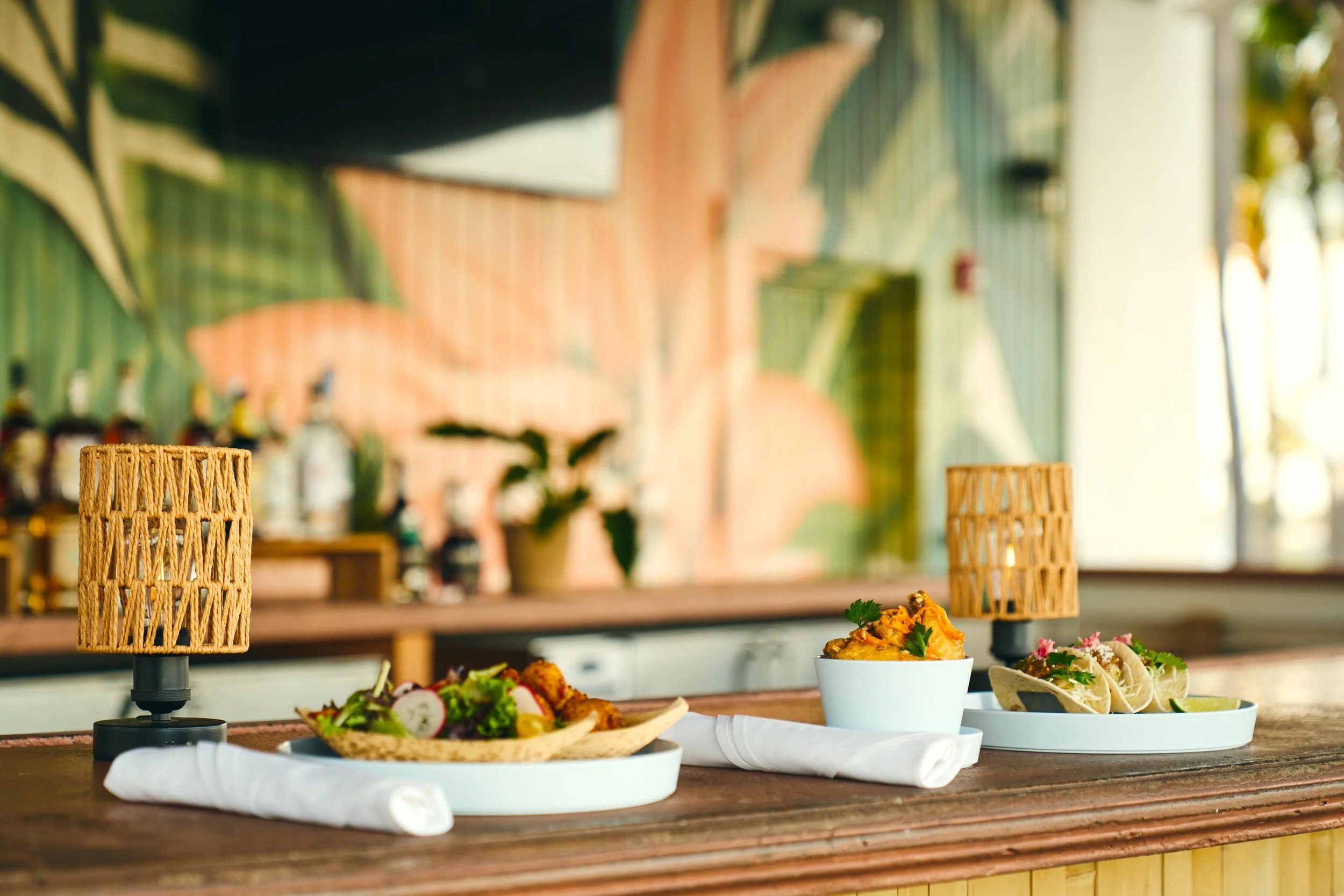 A close-up of a wooden table with three plates of food, two small woven lamps, and cloth napkins, inside a restaurant with tropical-inspired decor.