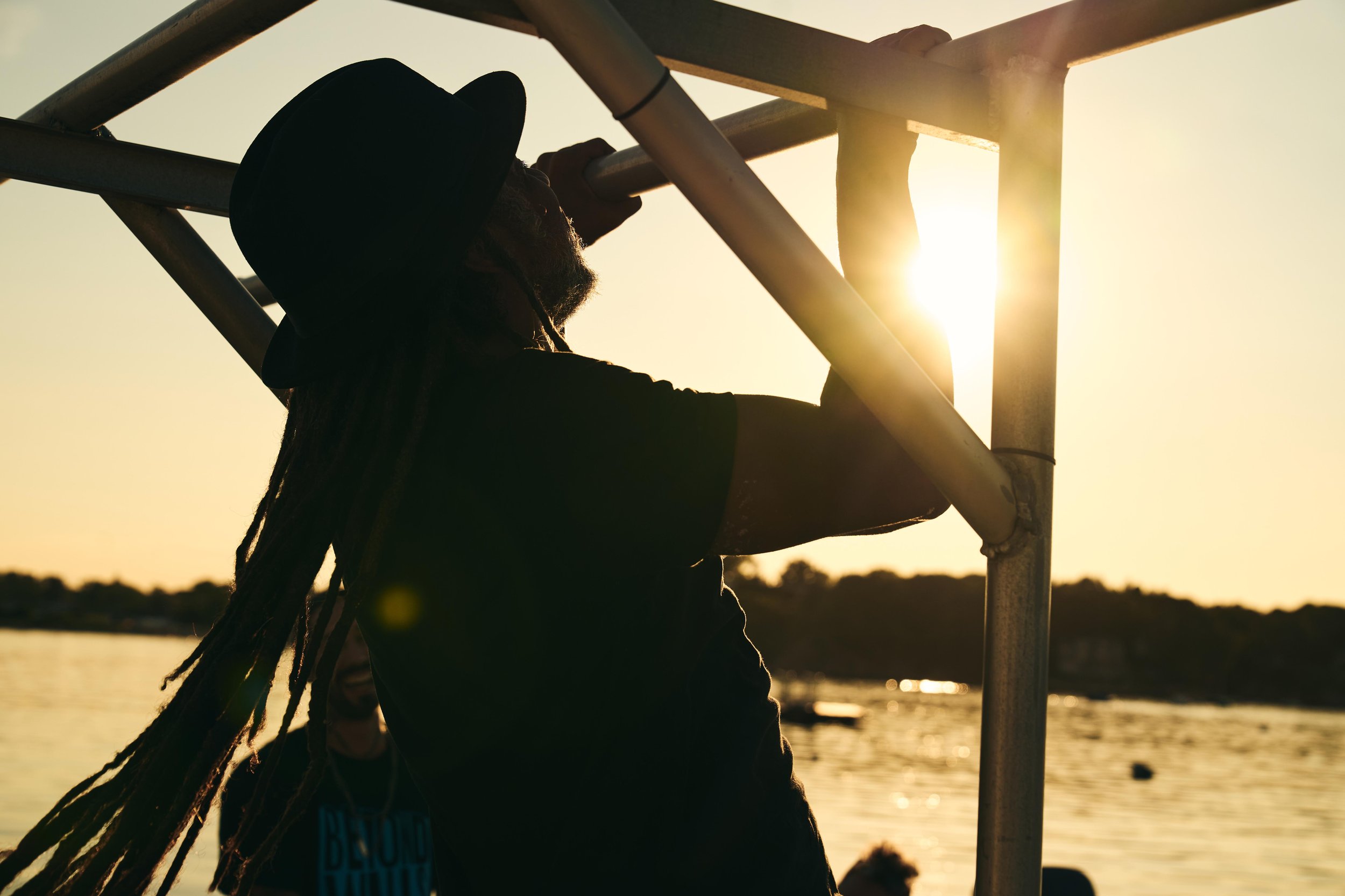 A person with long dreadlocks and a wide-brim hat climbing a metal structure with the setting sun in the background over a body of water.