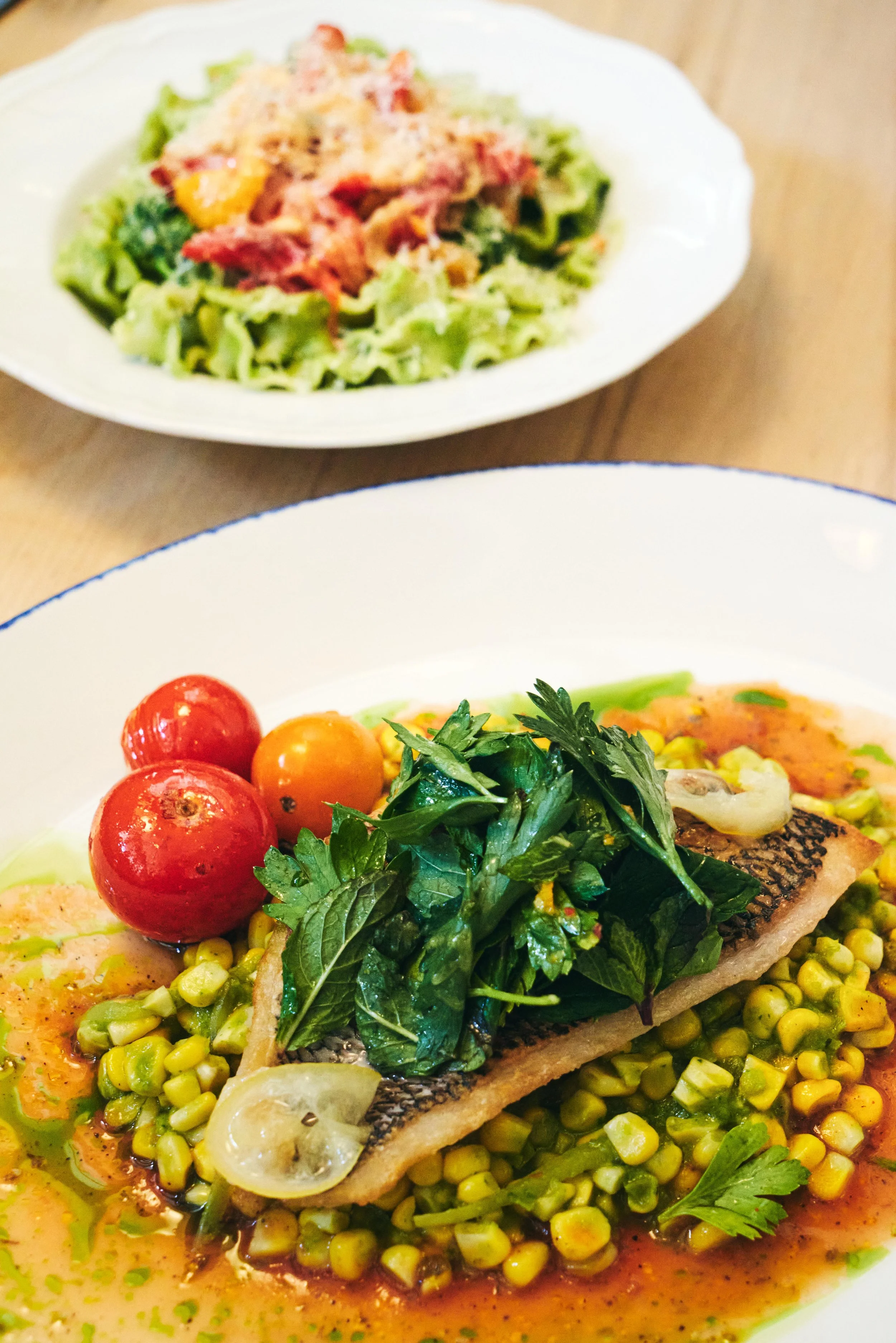 A plate of grilled fish topped with herbs and lemon slices, served with cherry tomatoes and corn on a bed of sauce, with a side salad of lettuce and shredded vegetables in the background.
