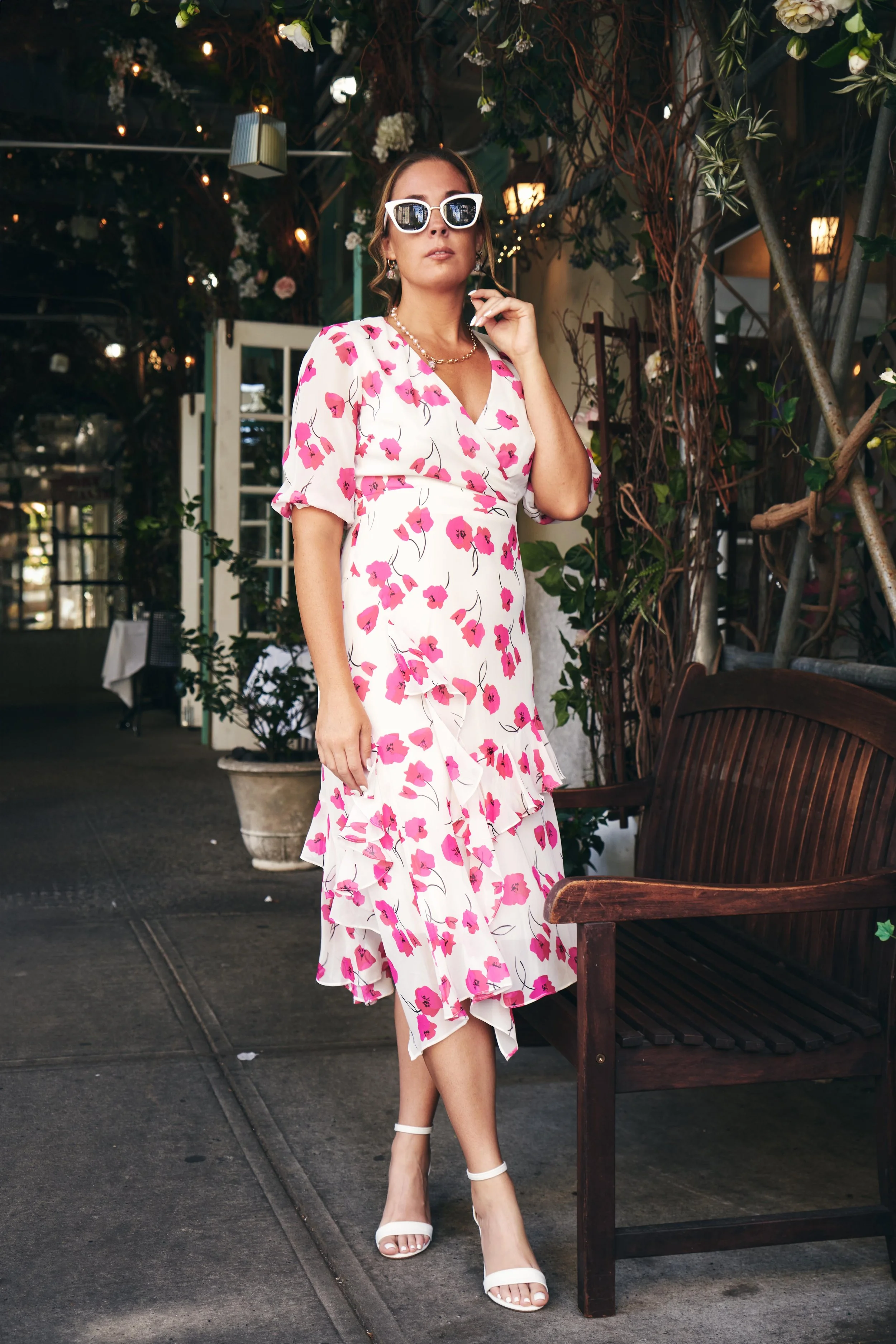 A woman in a white dress with pink floral pattern poses inside a restaurant or cafe. She is wearing white high-heeled sandals, white sunglasses, and jewelry, with one hand near her face and the other hanging by her side. The background includes plant