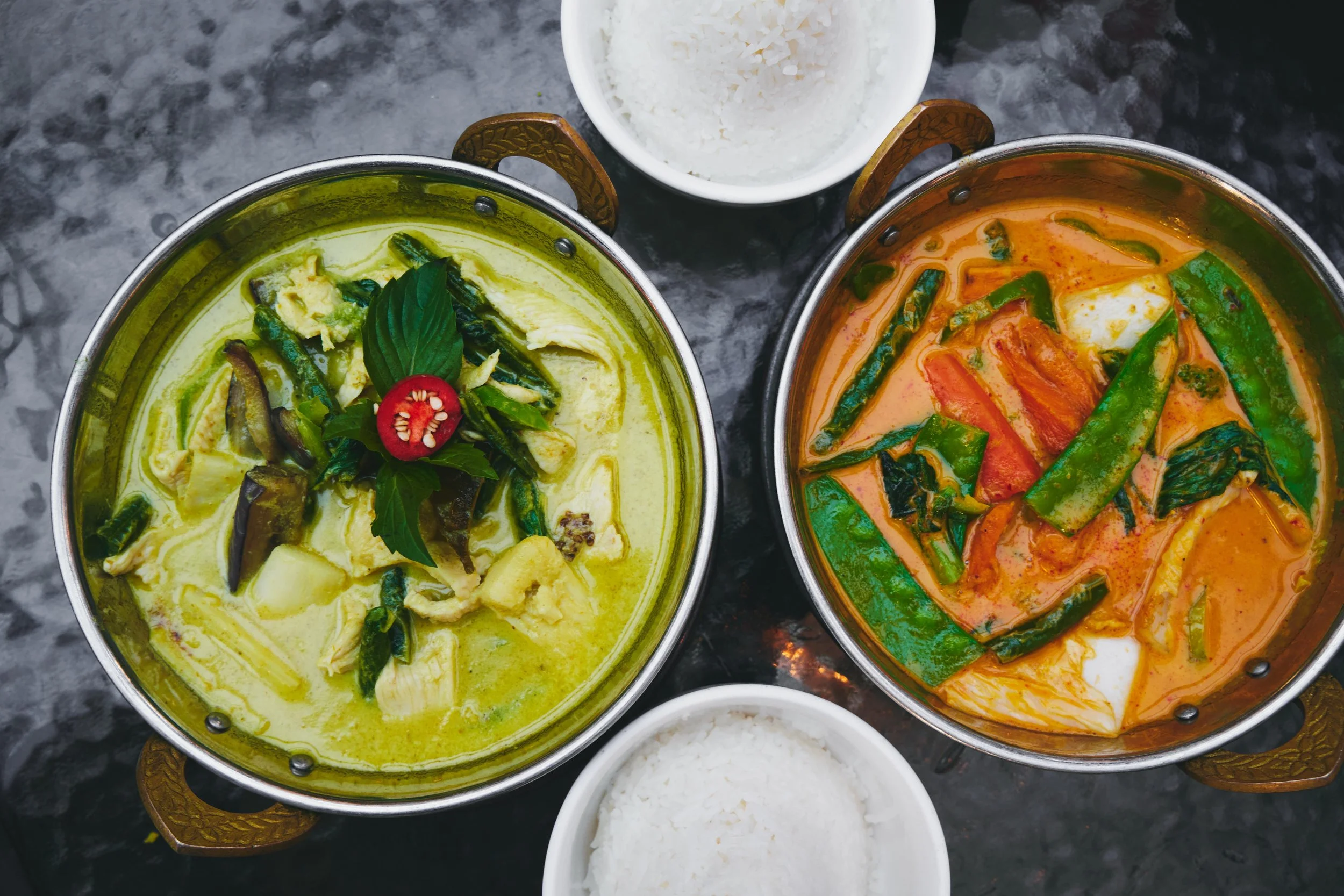 Two bowls of Thai curry, one green and one red, with fresh vegetables, basil, and red chili on top, accompanied by small bowls of white rice.