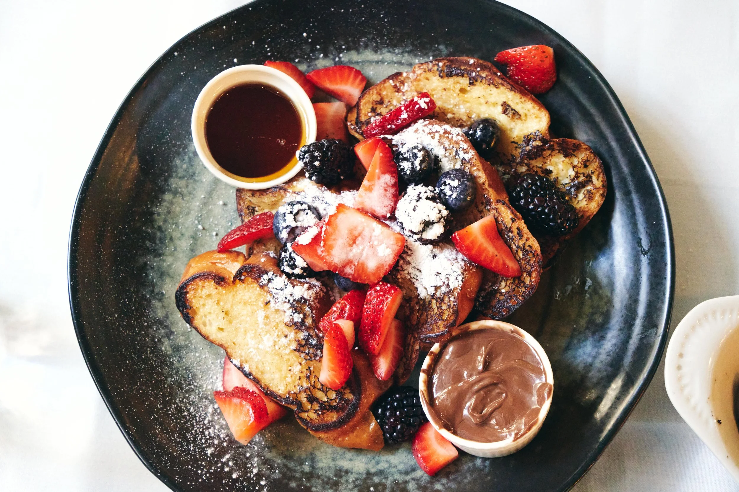 French toast topped with powdered sugar, strawberries, blueberries, blackberries, and raspberries, served with chocolate spread, syrup, and additional syrup on the side.