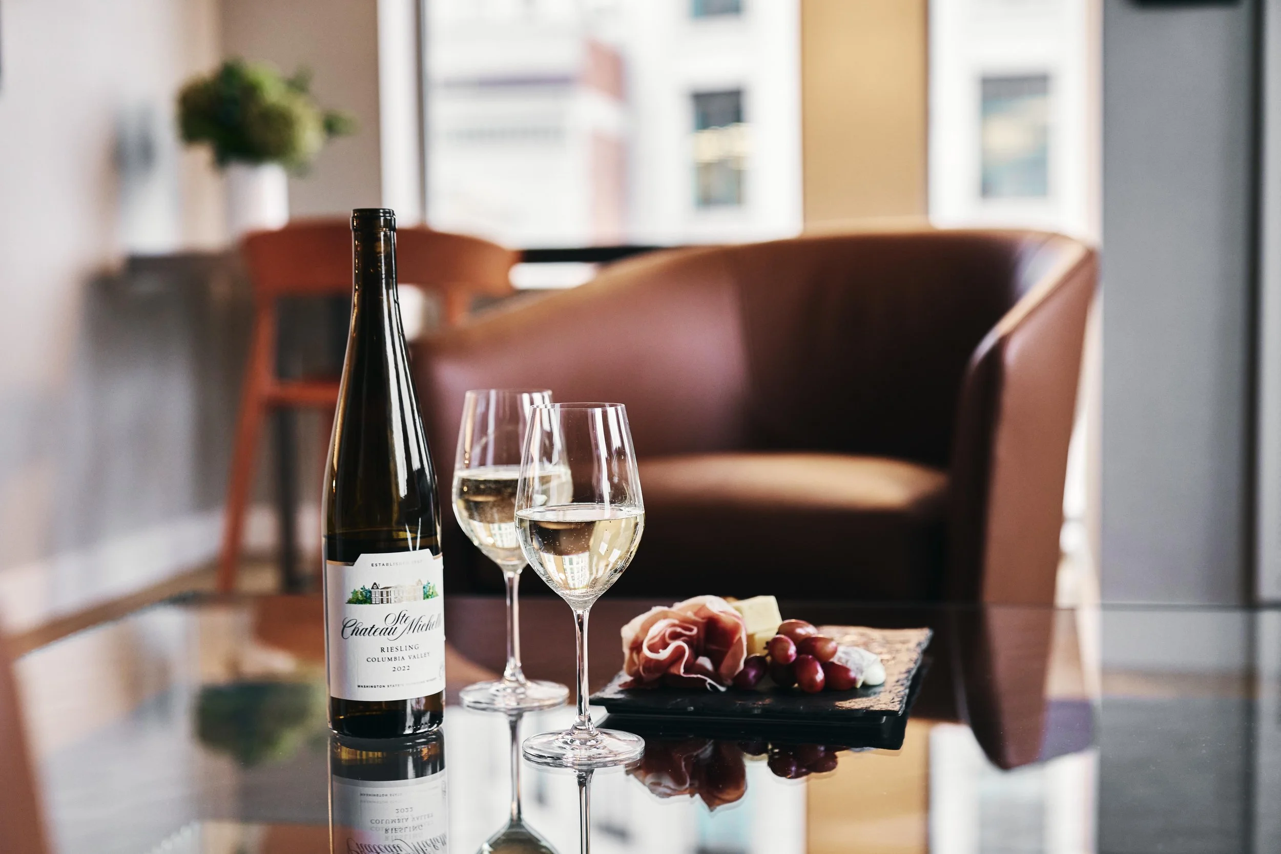 A bottle of Chardonnay wine, two glasses of white wine, and a platter of cheese, grapes, and meat on a glass table in a cozy room with a brown leather sofa and large windows.