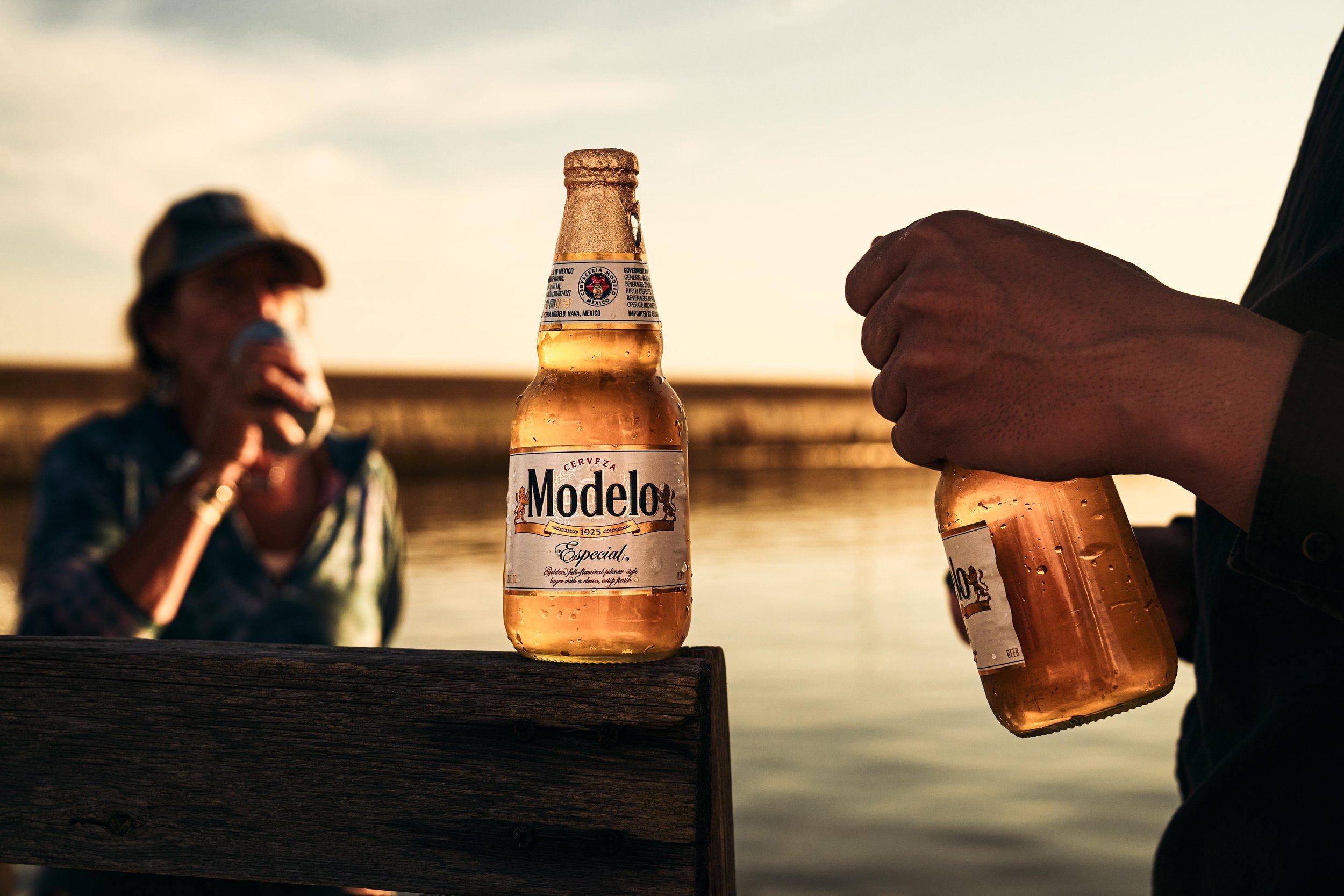 Two people sharing Modelo beer bottles by the water at sunset, with a woman in a hat drinking from a can in the background.