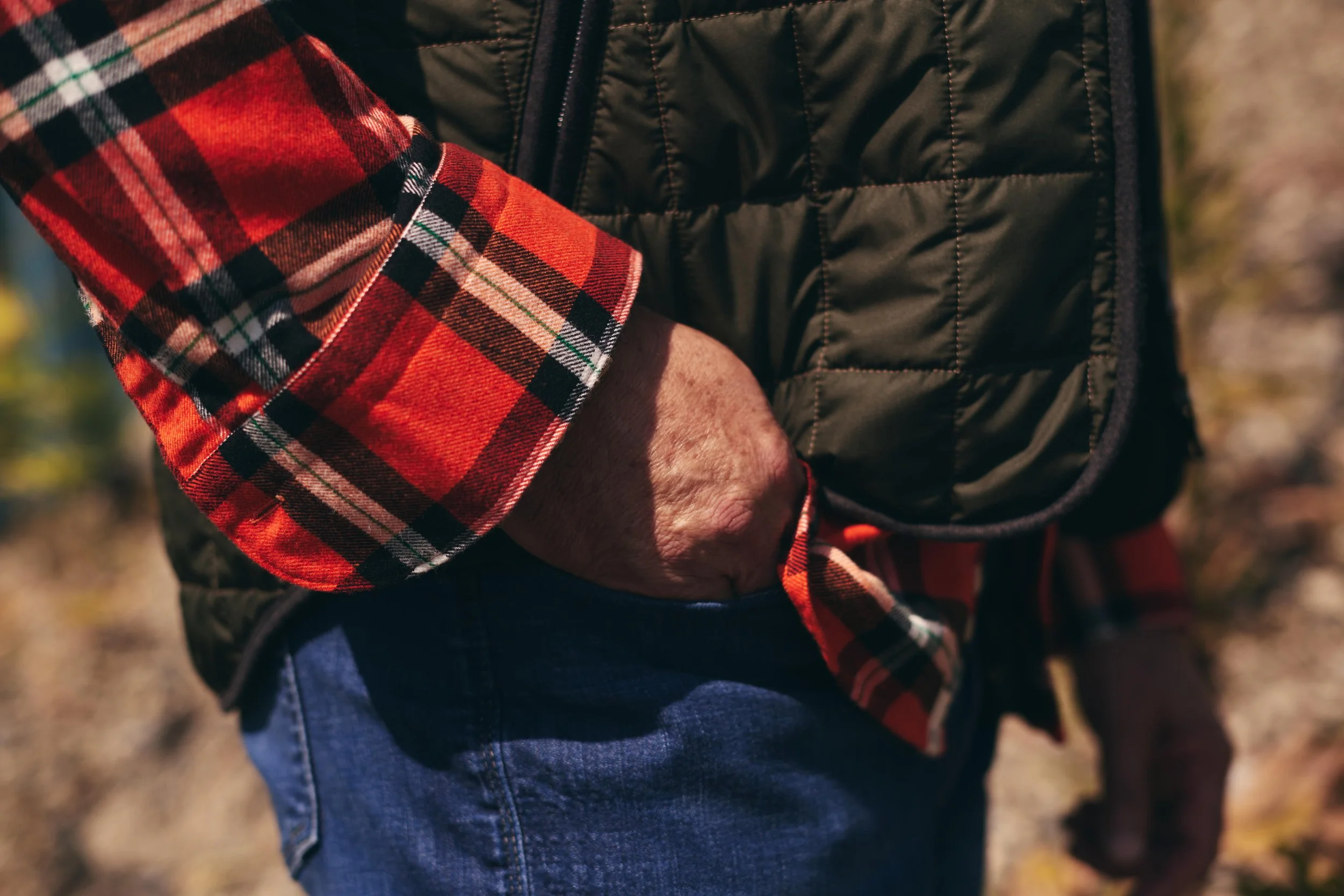 Close-up of a person's hand in their pocket, wearing a red and black plaid shirt with a quilted black vest and blue jeans, outdoors.