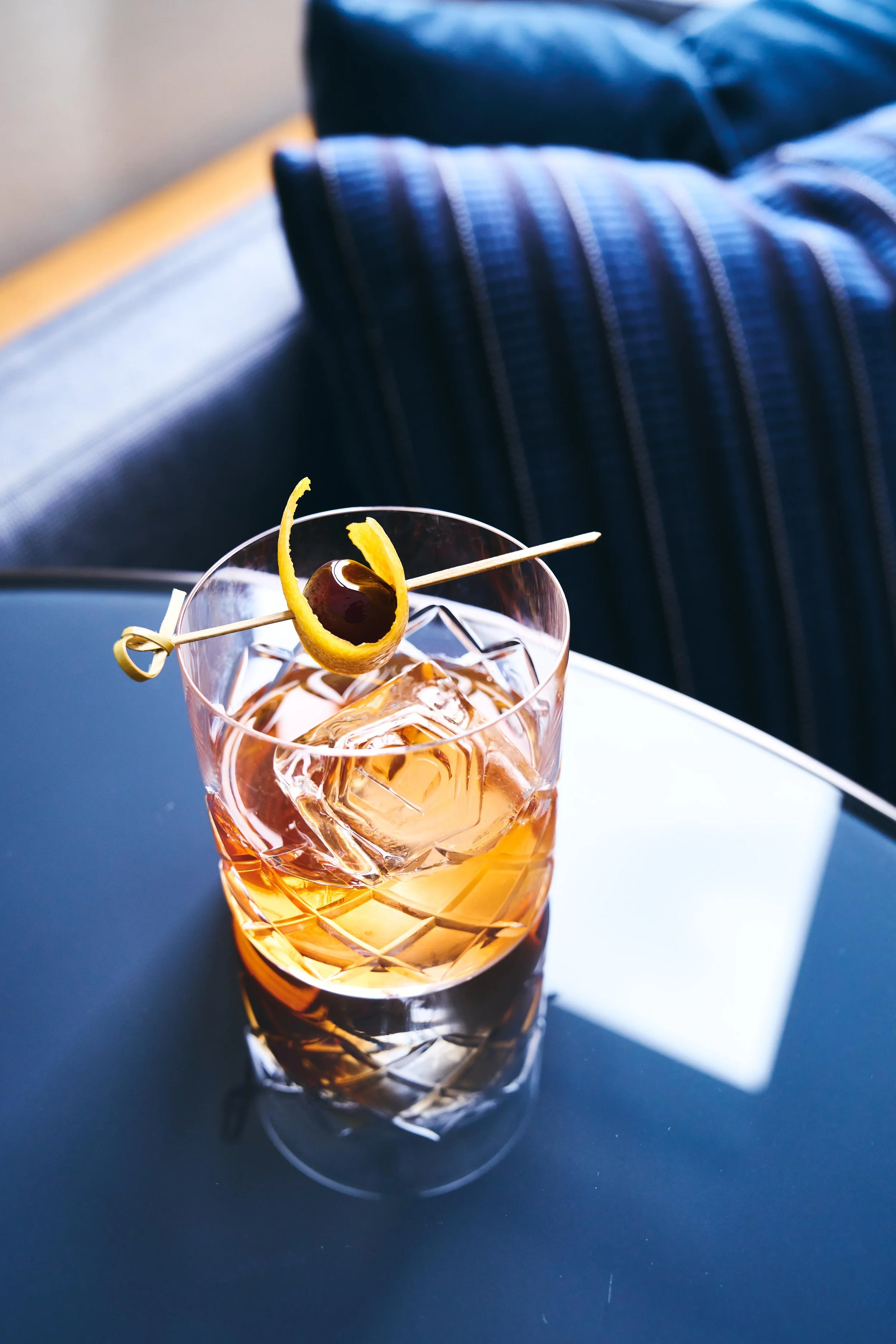 A glass of amber-colored cocktail with ice cubes, garnished with a lemon peel and cherry skewer, on a dark table.