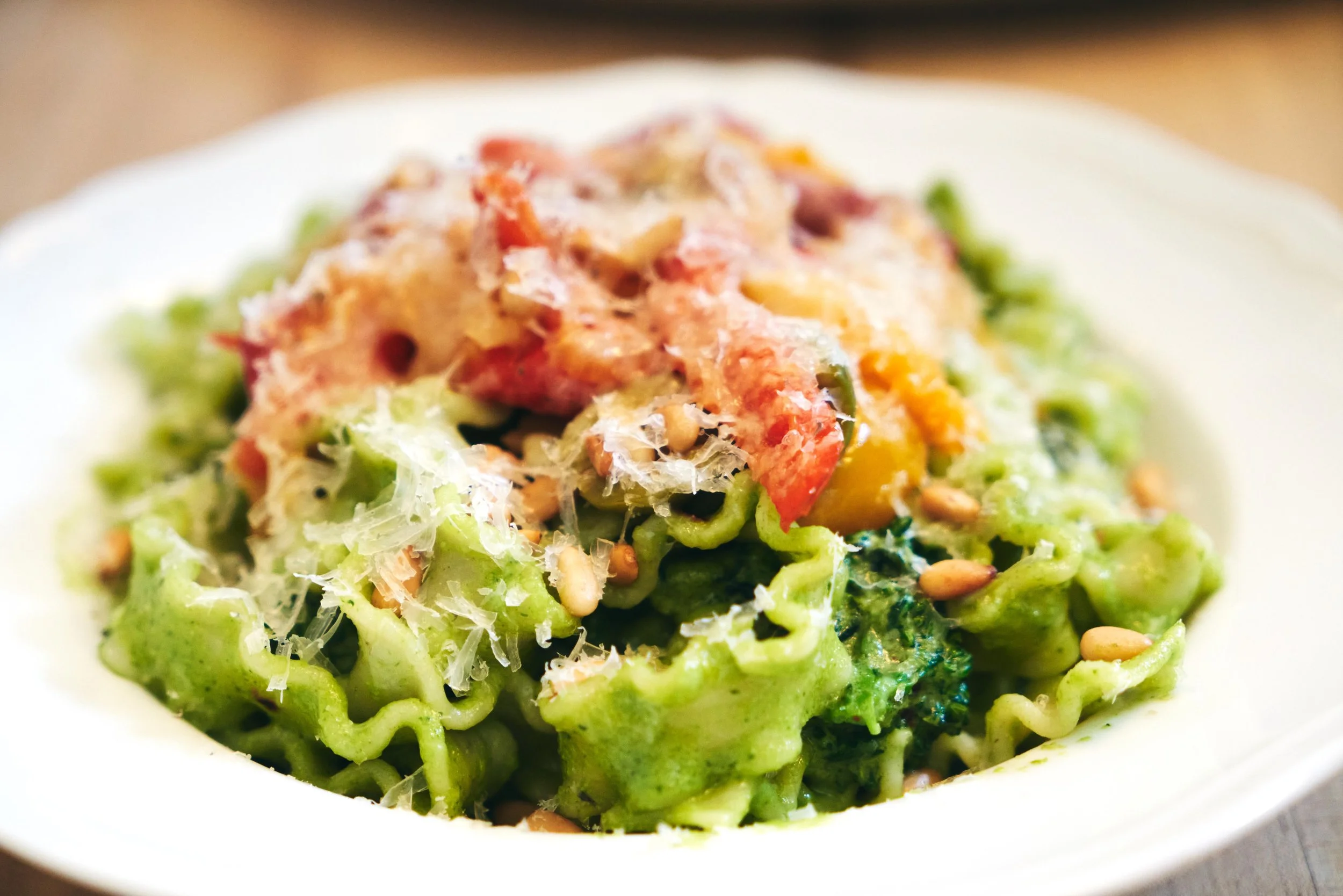 Bowl of cooked pasta with green sauce, topped with grated cheese, tomatoes, and sunflower seeds