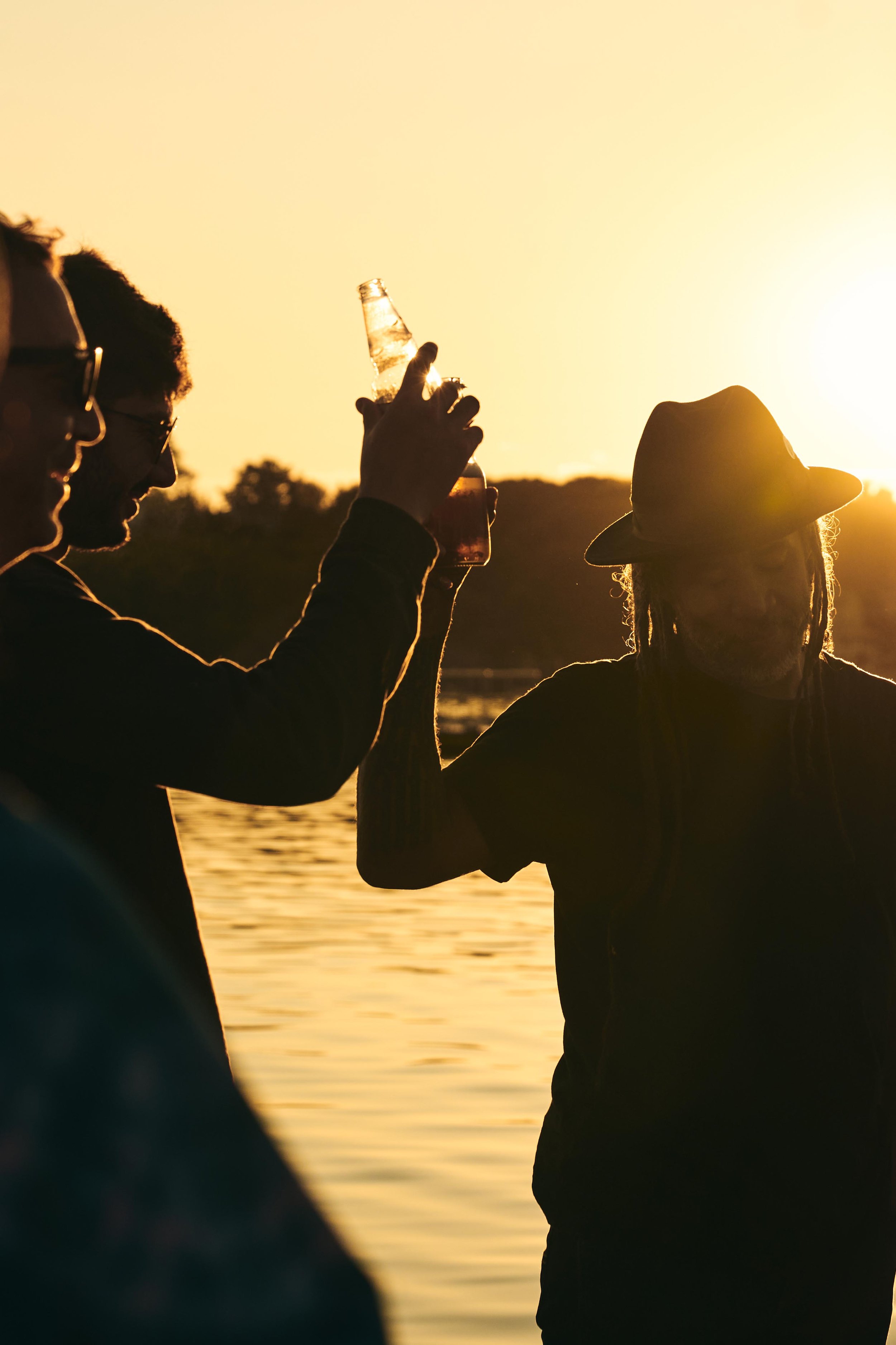 People silhouetted against sunset by the water, one holding a beer bottle and another raising a glass.