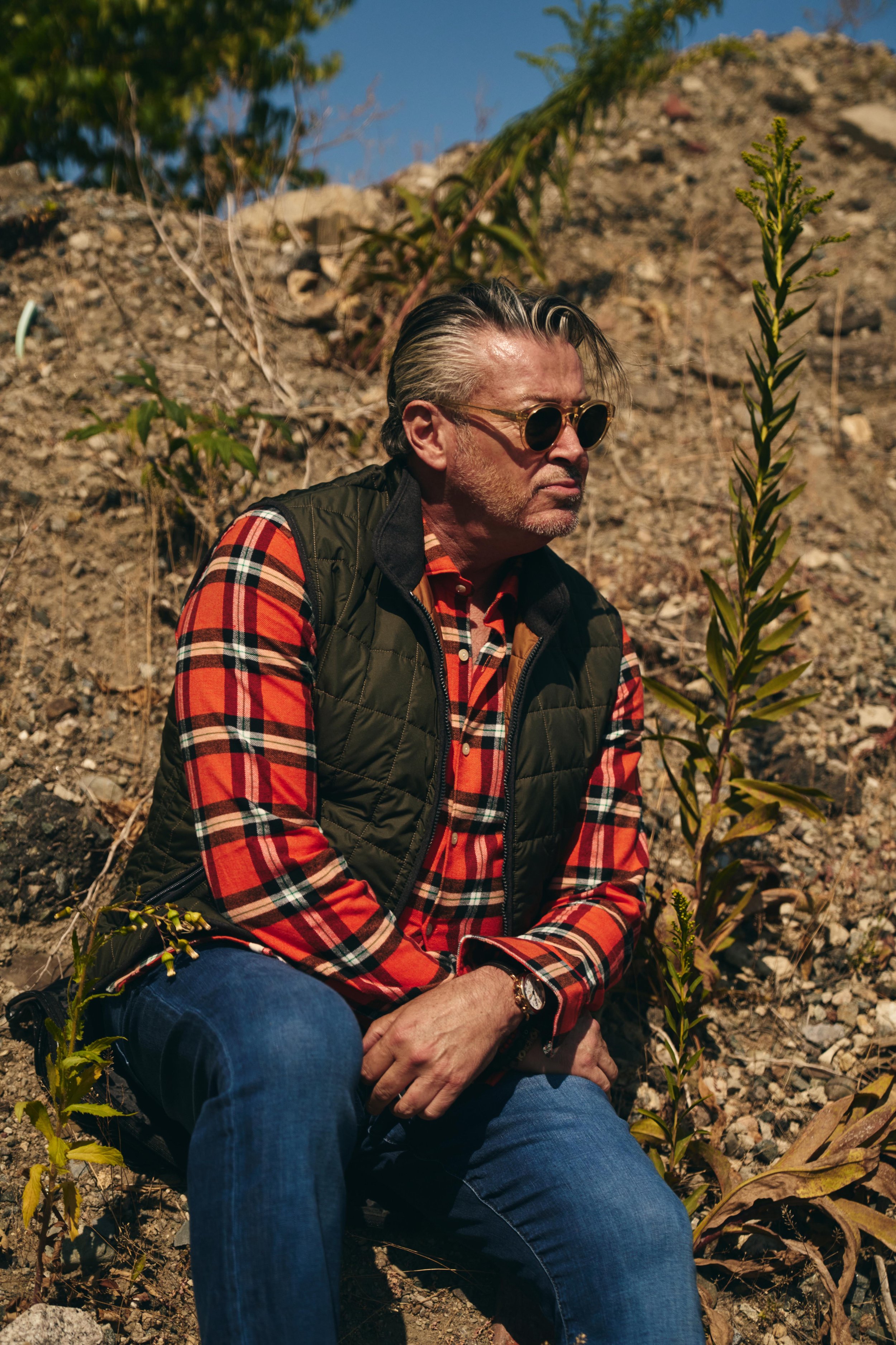 A middle-aged man with sunglasses, wearing a red plaid shirt and a dark vest, sitting on rocky ground outdoors with sparse plants and a blue sky in the background.