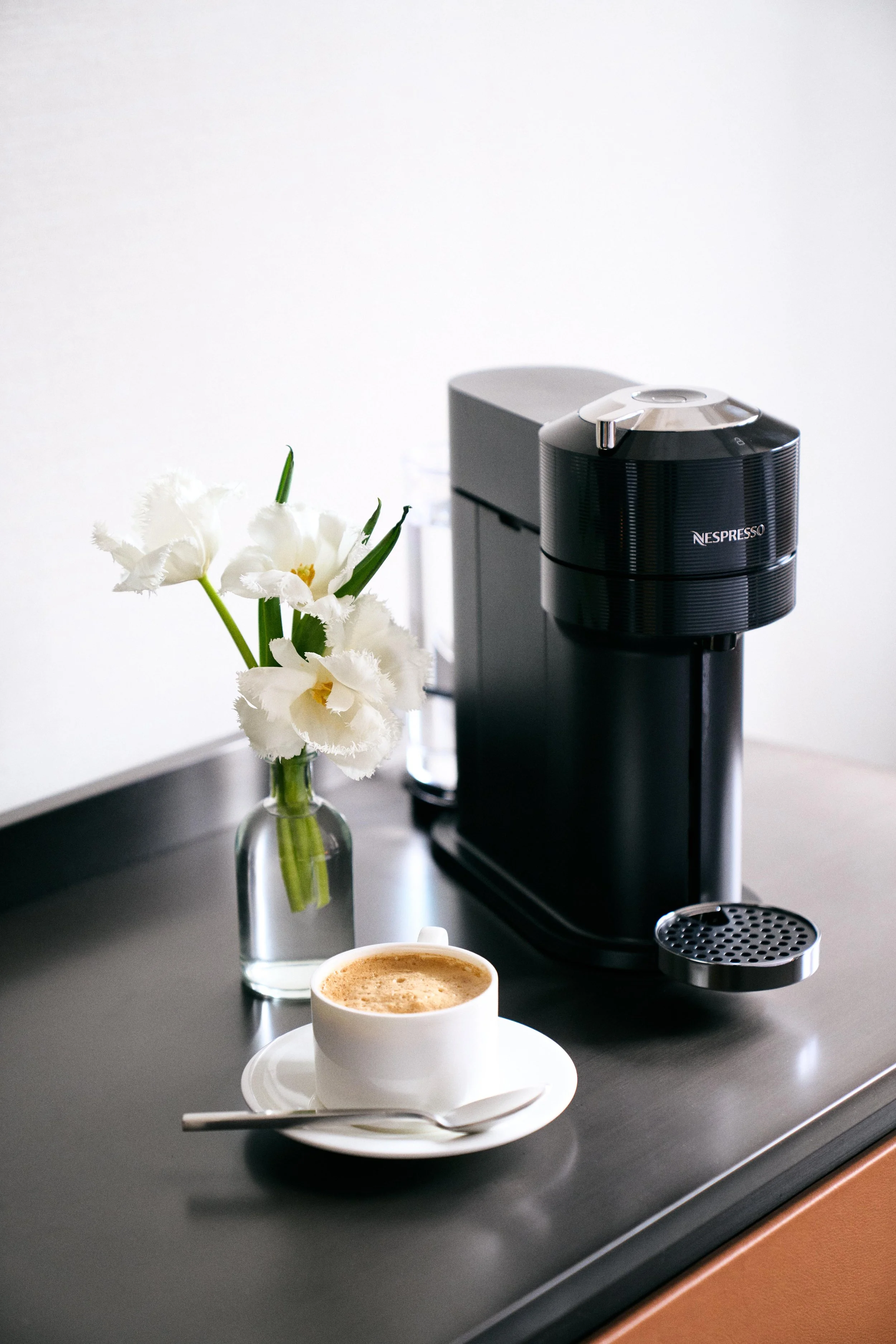A black Nespresso coffee machine, a white cup of coffee on a saucer with a spoon, a small glass bottle of white flowers, and a black countertop.