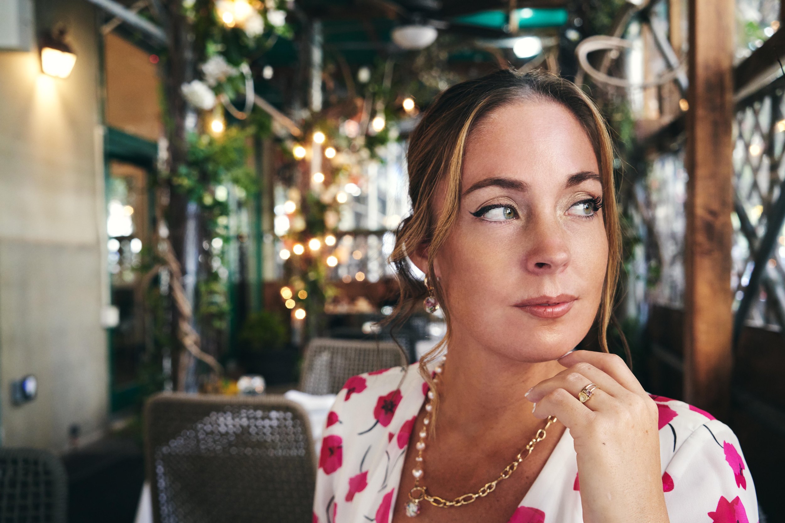 A woman with light brown hair in a loose updo, wearing a white blouse with pink floral print, gold jewelry, and makeup with eyeliner, sitting in a warmly lit indoor setting with greenery and string lights in the background, looking to her right with 