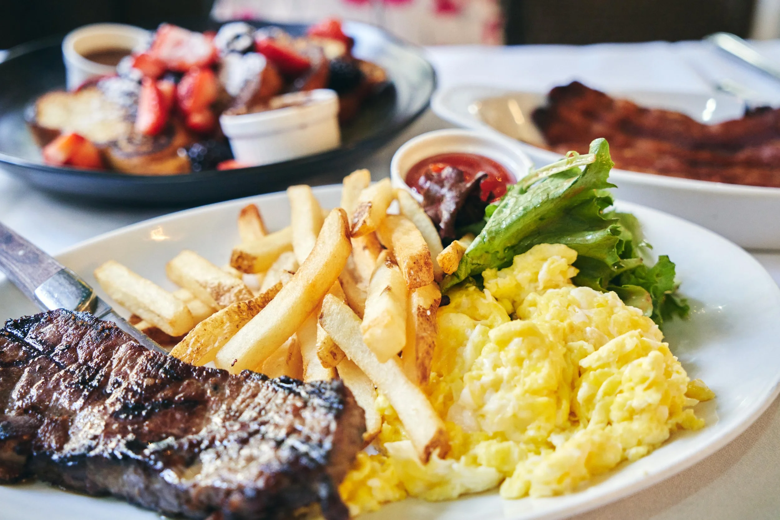 Breakfast plate with scrambled eggs, French fries, salad greens, and grilled steak, with side dishes of toast, bacon, strawberries, and syrup in the background.