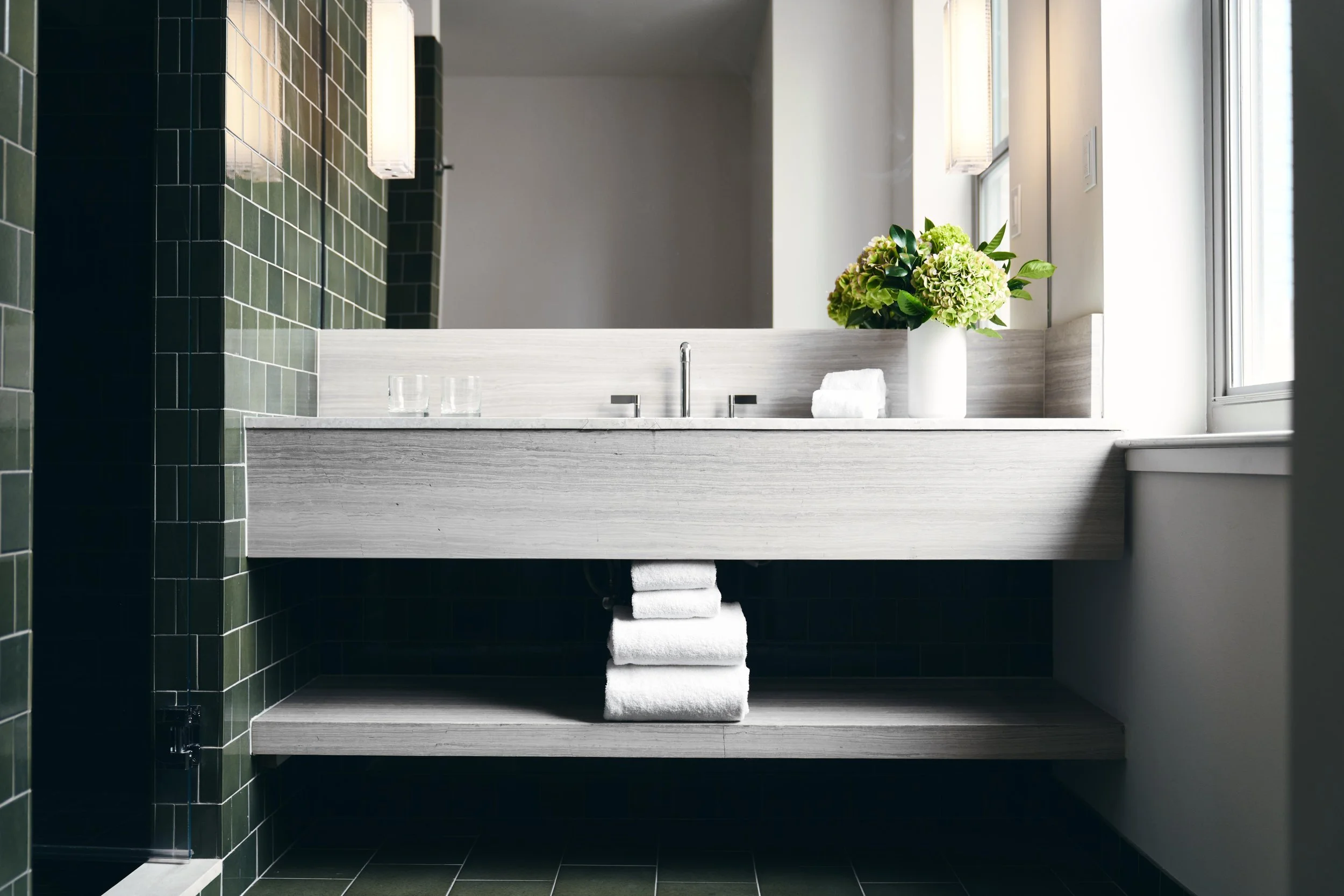 Modern bathroom with a white floating vanity, a large mirror, green tiles on the wall, and a window with natural light. A vase with green flowers and folded towels are on the vanity.