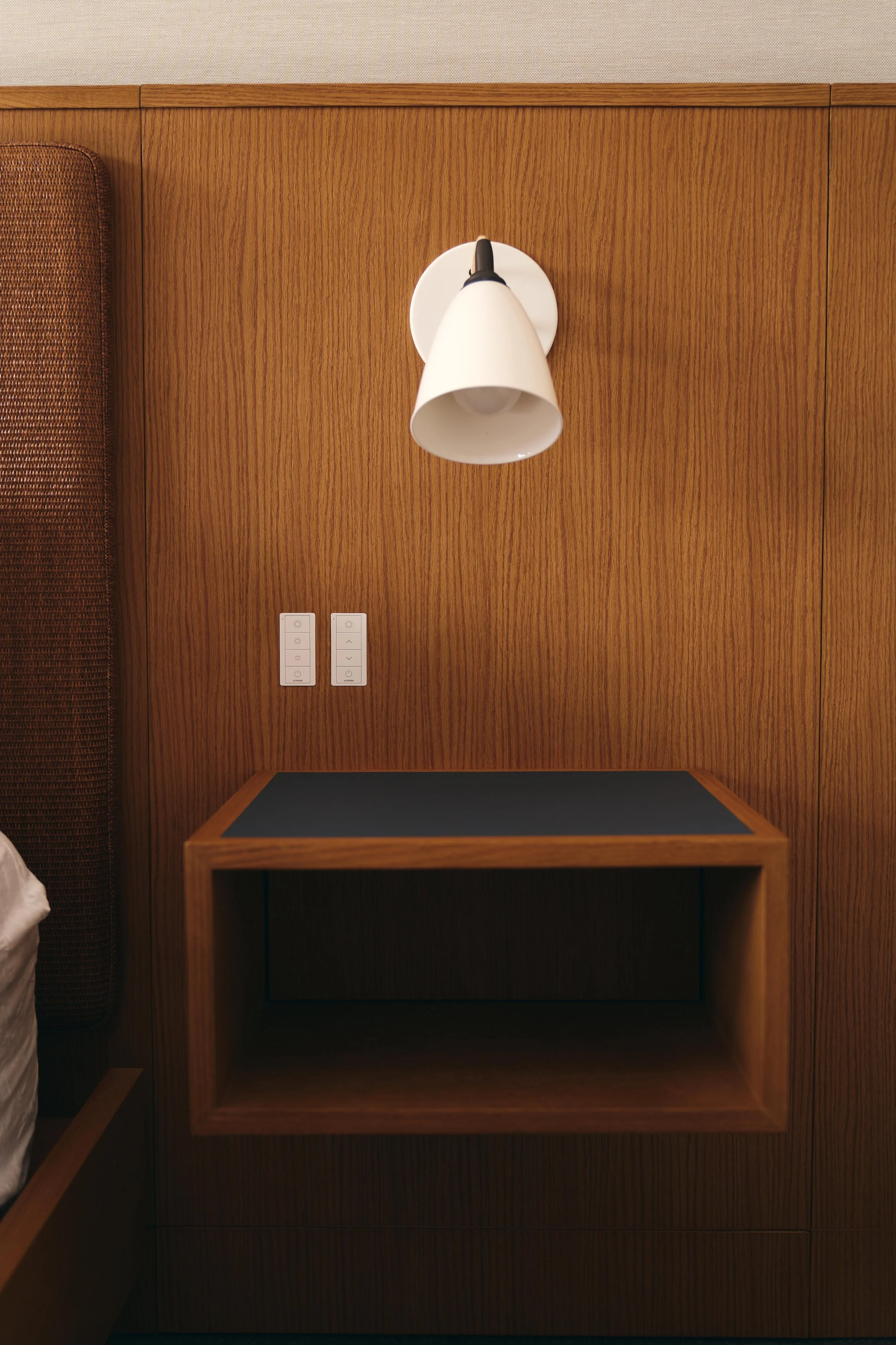 A wooden headboard with a white wall-mounted lamp and two switches on a wood-paneled wall.