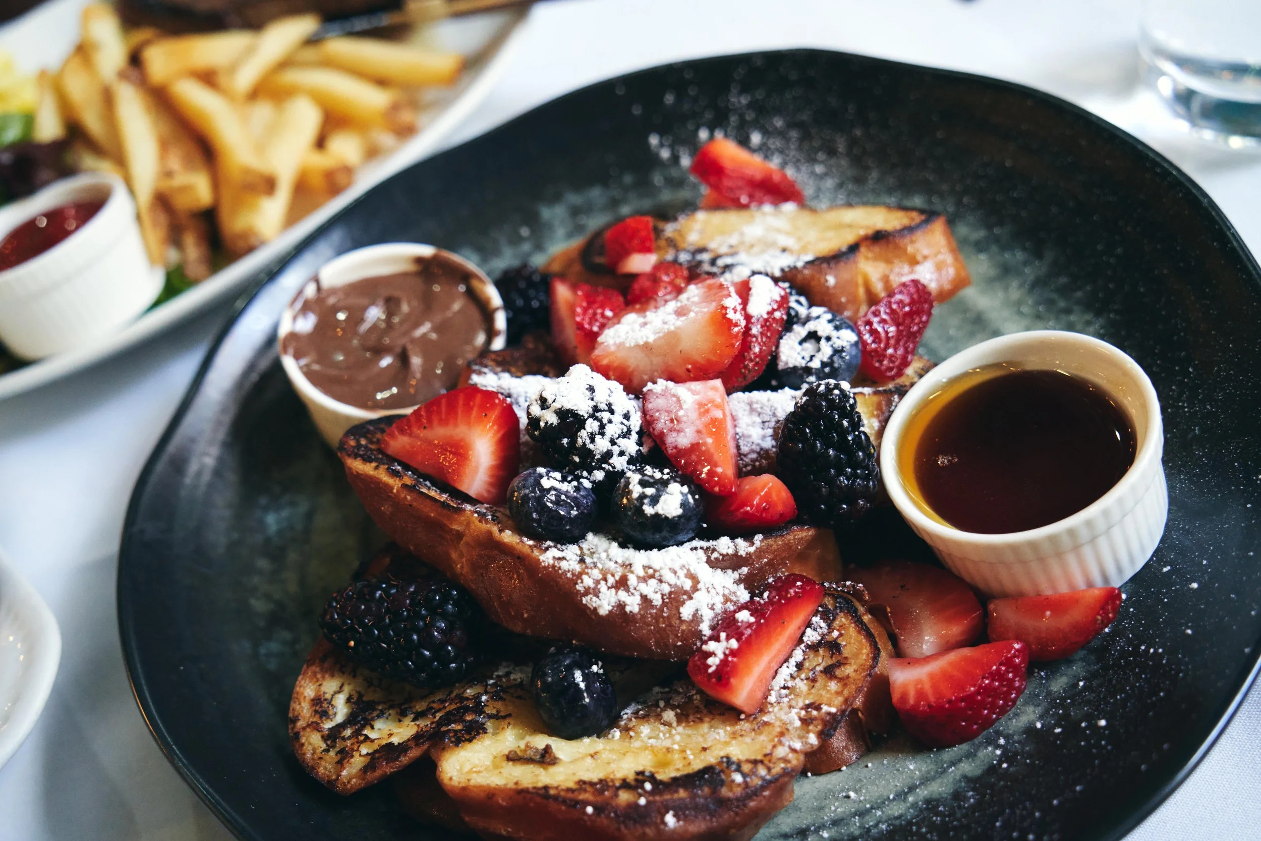 French toast topped with fresh strawberries, blackberries, blueberries, and powdered sugar, served with syrup and chocolate sauce.