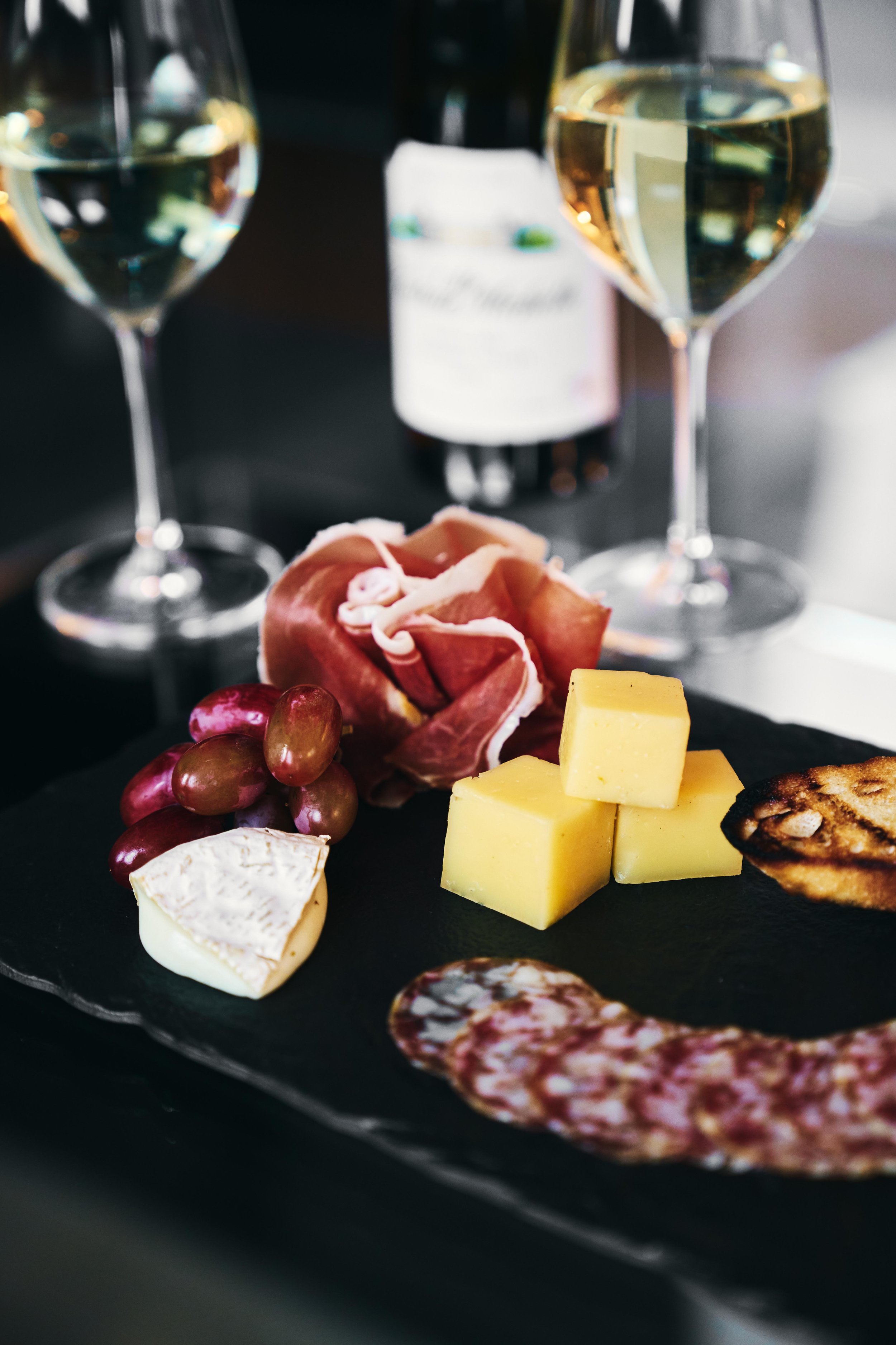 A charcuterie board with cheeses, grapes, slices of cured meat, and crackers, accompanied by two glasses of white wine and a wine bottle in the background.